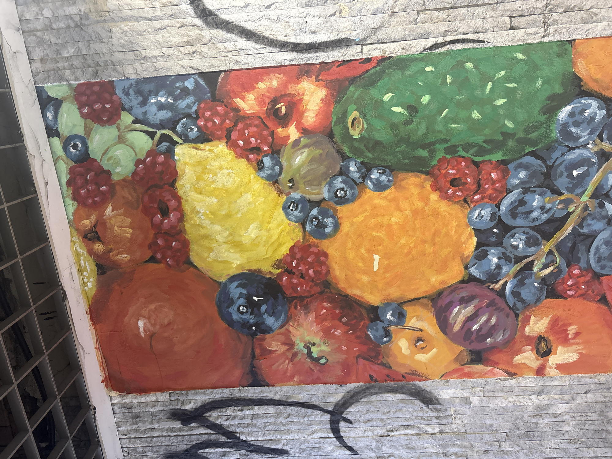 A rectangular painting of assorted fruits on a wall composed of horizontal stacked stone blocks. The fruits depicted include blackberries, raspberries, blueberries, grapes, pineapple, apples, peaches, plums, and an orange. The location is Unknown, Unknown. The painting is mounted on the textured stone wall. There is graffiti at the top and bottom of the painting.