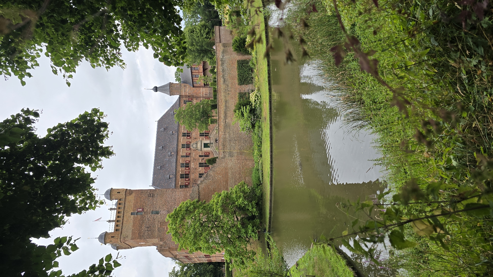 The image depicts a view of a brick castle, identified as Bergh Castle in 's-Heerenberg, Netherlands, surrounded by a moat. The castle, constructed from red brick, features multiple towers, including a prominent square tower with battlements and two smaller, domed turrets on its left. Adjacent to this tower is a large, pitched-roof building with numerous windows. A smaller, conical-roofed tower is visible further to the right. The castle is partially obscured by lush green foliage from trees in the foreground and framing the top of the image. The moat, filled with murky water, reflects the castle and the overcast sky. The grounds around the castle are manicured with green lawns and low hedges. The overall scene is serene and picturesque, with no people or animals clearly visible.