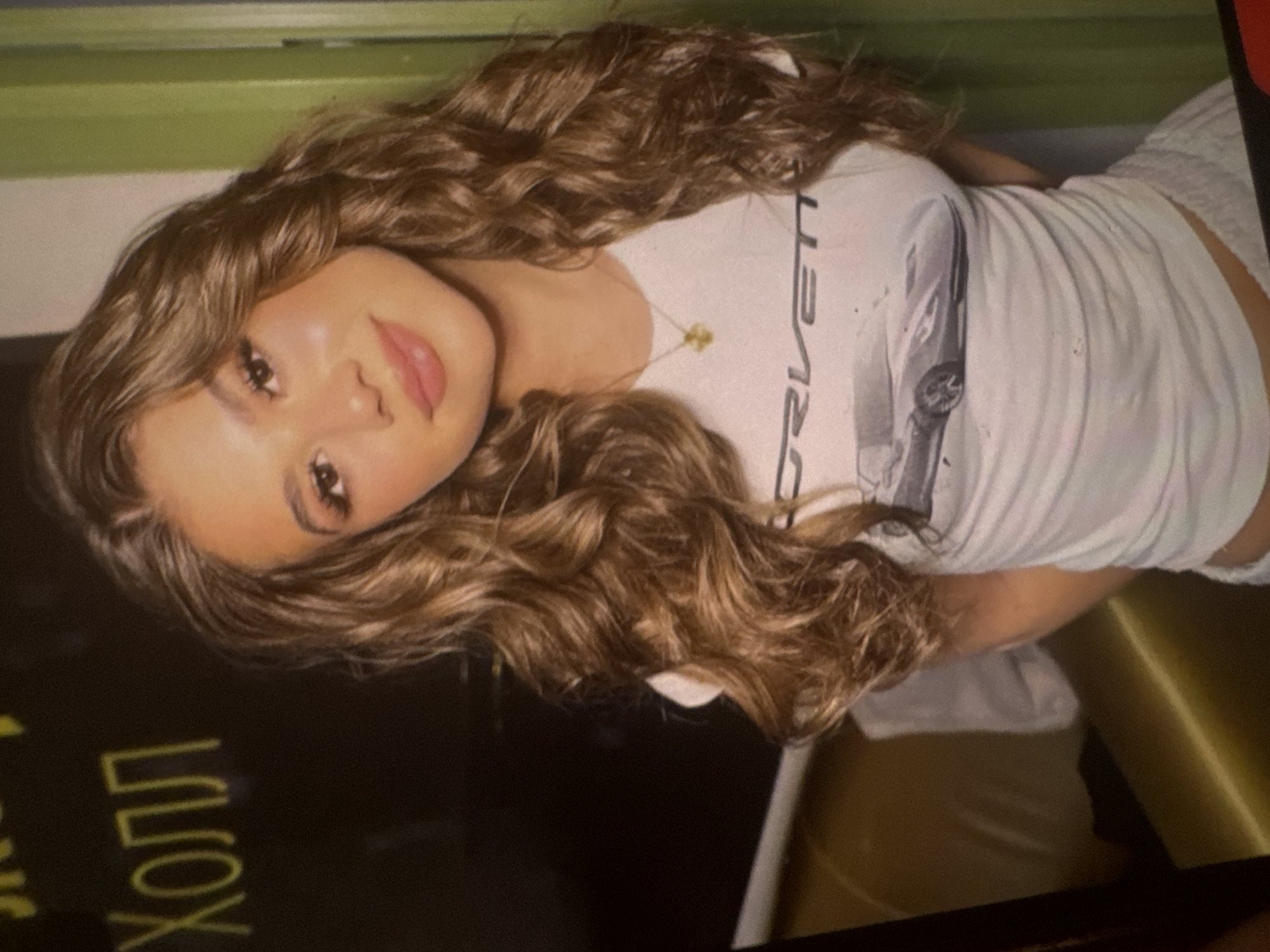 An indoor, sideways-oriented photograph captures a young woman posing for the camera with a soft, pleasant expression. She has long, wavy, light brown hair and fair skin, with noticeable eye makeup including winged eyeliner and pink lipstick. She is wearing a white, fitted, short-sleeved t-shirt that features the word "DRIVEN" and a graphic of a car, with additional letters "RLett" visible below the car, possibly indicating "Corvette" or a related car brand. A delicate, light-colored necklace with a small pendant is visible on her chest.
The background consists of a pale green wall or surface behind her head. To her side and below her, there is a dark, indeterminate surface, and a reflective, gold or bronze-colored surface, possibly indicating furniture like a booth or seat upholstery. In the foreground, on a dark surface, yellow Cyrillic text is visible, spelling "ХОЛЛ," which translates to "Hall" in Russian, suggesting the location could be a hall, lobby, or similar interior space. The lighting appears artificial, and the image has a slightly grainy quality.