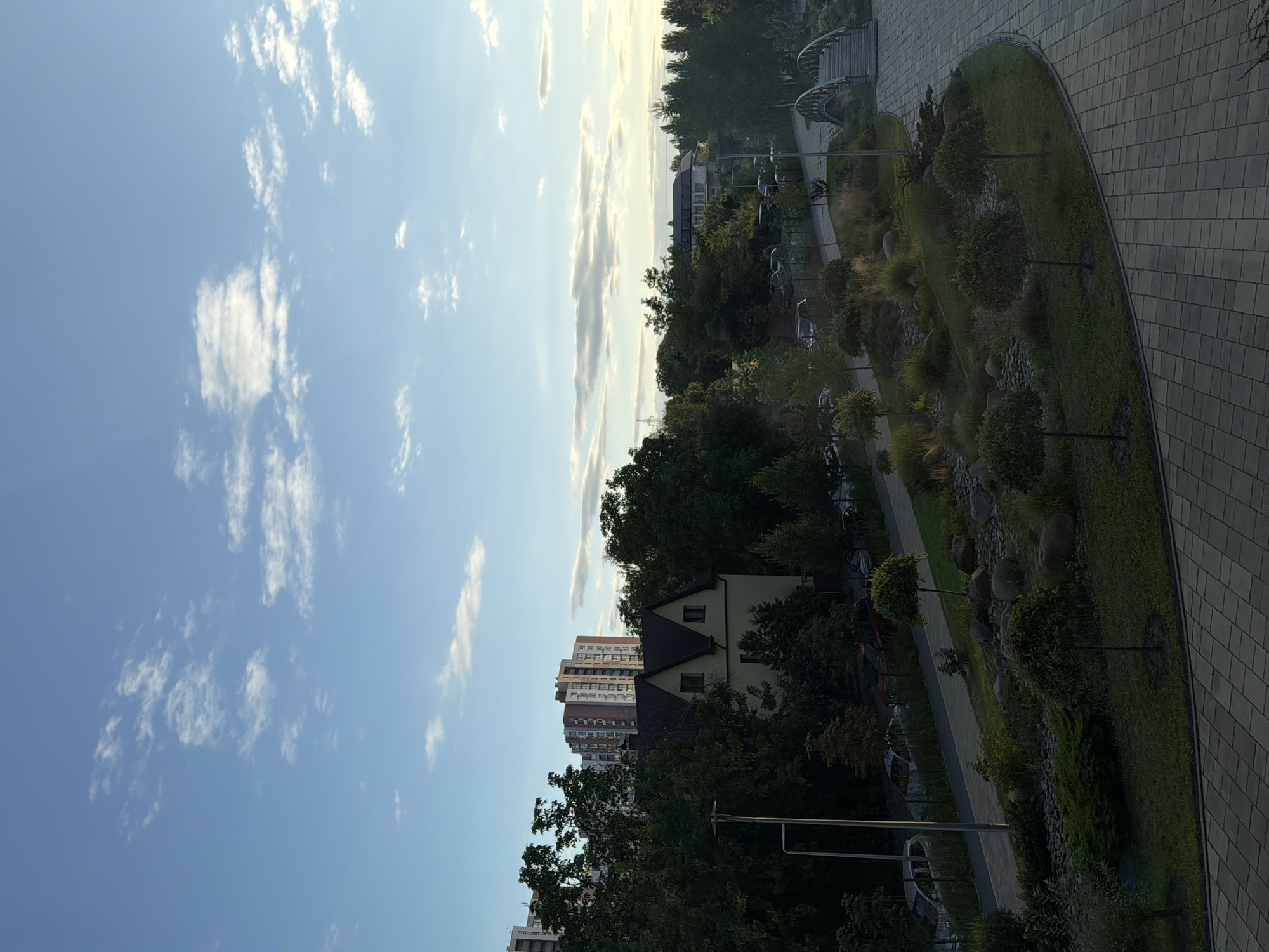 The image shows a park area with a paved walkway and a decorative garden. In the background, there are trees, residential buildings, and a small bridge. The sky is blue with scattered clouds. The scene appears to be an outdoor urban environment. The perspective suggests the photo was taken from an elevated position. The lighting indicates it is likely daytime, possibly late afternoon due to the soft light.