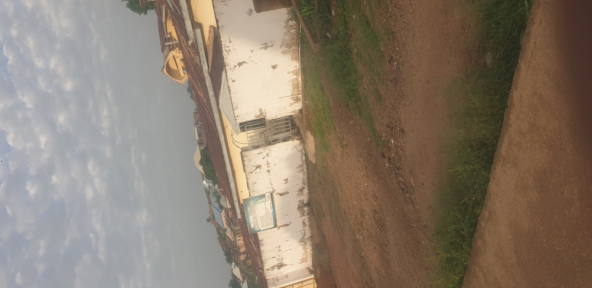 The image shows the exterior of the "PRO 11 (NASARA) SPECIALIST HOSPITAL" in Jos, Nigeria. A weathered, white wall with peeling paint surrounds the property. On the wall, a rectangular sign displays hospital services, including "Specialist Clinics," "Consulting Clinics," "Diagnostic Services," and "Surgical Procedures." A metal gate, partially open, leads into the hospital grounds. In the background, various buildings with corrugated iron roofs are visible, some with arched architectural features, suggesting a mix of residential and commercial structures common in the city. The sky above is partly cloudy, with a hazy, muted light, indicating it might be early morning or late afternoon. The foreground consists of a dirt path and patches of green grass, typical of the landscape in Jos. There are no people visible in the frame.