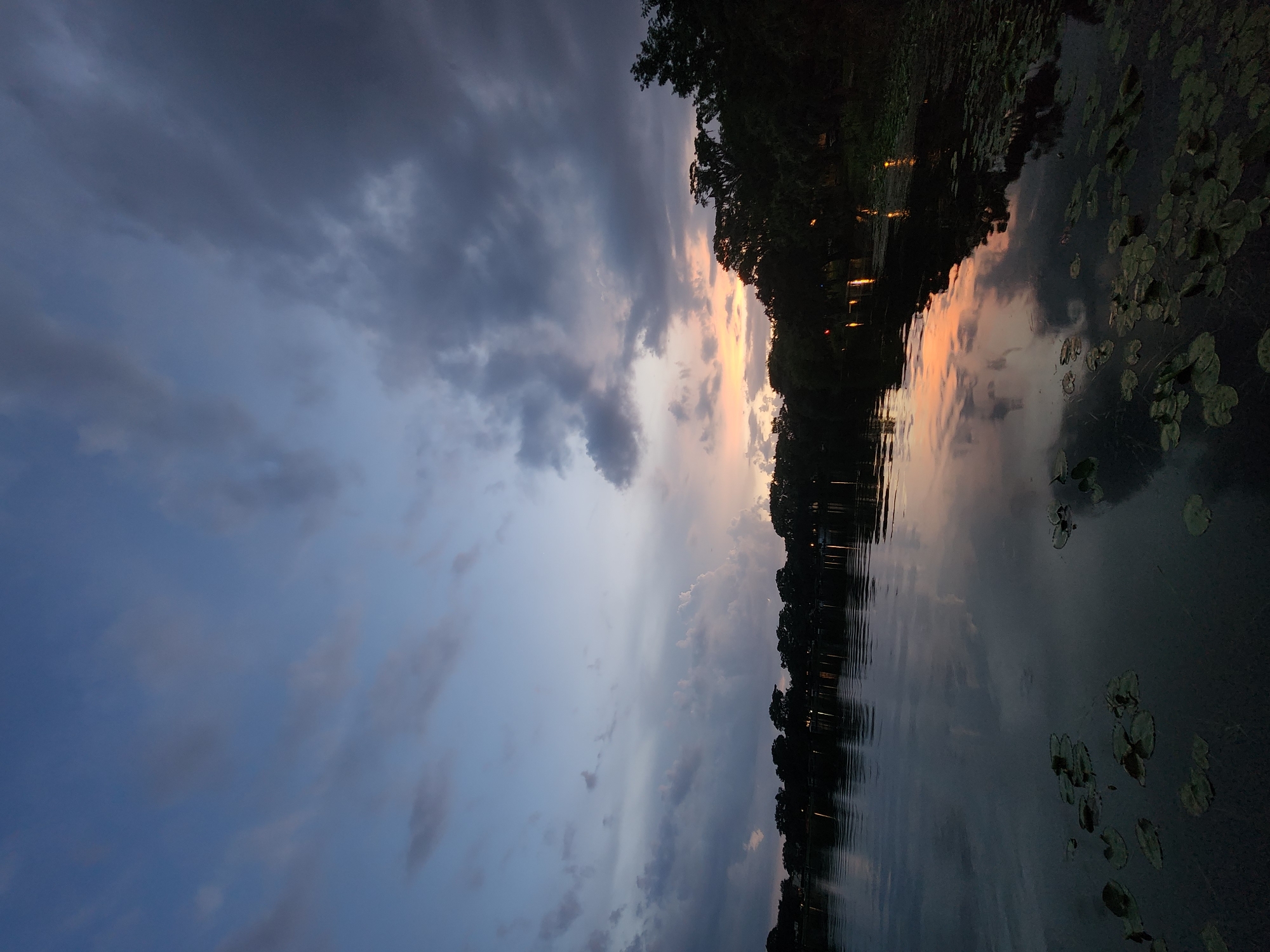 The image depicts a serene lakeside scene at dusk in Winter Park, Florida. The sky is a gradient of blues, grays, and soft oranges, with scattered clouds. The calm water of the lake reflects the sky and the silhouettes of trees lining the distant shore. Lily pads are visible floating on the surface of the water in the foreground. The overall atmosphere is peaceful and tranquil, characteristic of twilight in a natural setting.