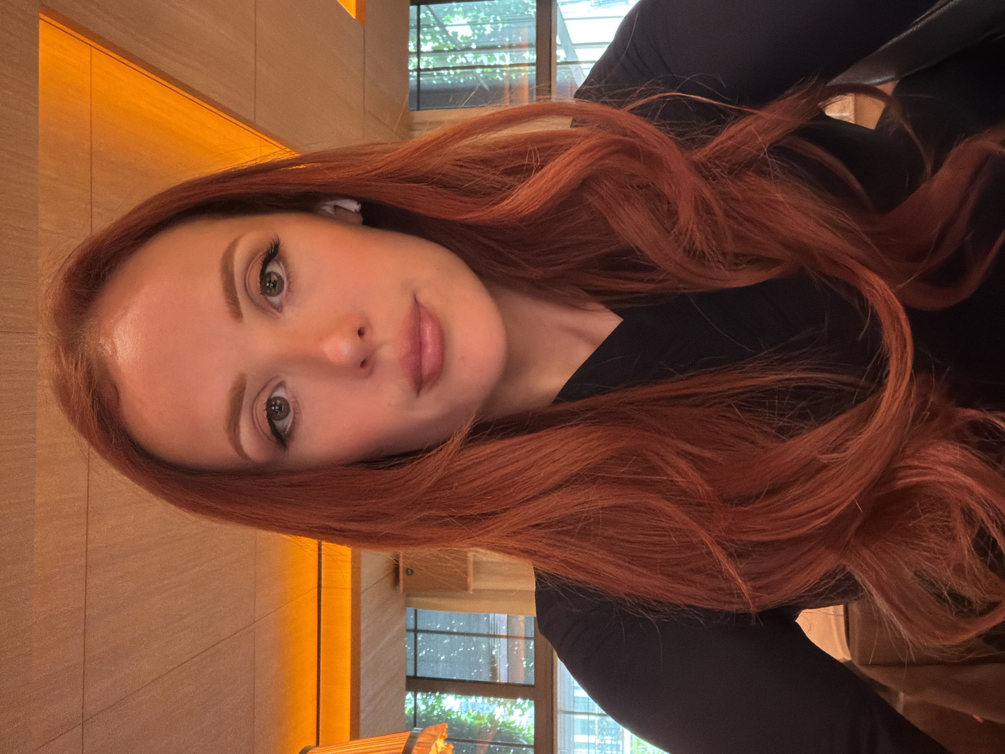 A woman with long, wavy, auburn hair is positioned in the foreground, angled slightly towards the viewer. She has fair skin, light eyebrows, and green eyes. She is wearing a black long-sleeved shirt. An in-ear headphone is visible in her left ear.
Behind the woman, a blurry cityscape with trees and high-rise buildings is visible through a window. The light suggests it's either morning or early evening. A wall with light colored, textured panels and a thin, orange light fixture runs along the left side of the frame.
The scene appears to be indoors, possibly in a hotel or restaurant. Although not definitively proven, the presence of the cityscape and window suggest the photo was taken in an urban location, but does not show it is Paris.
