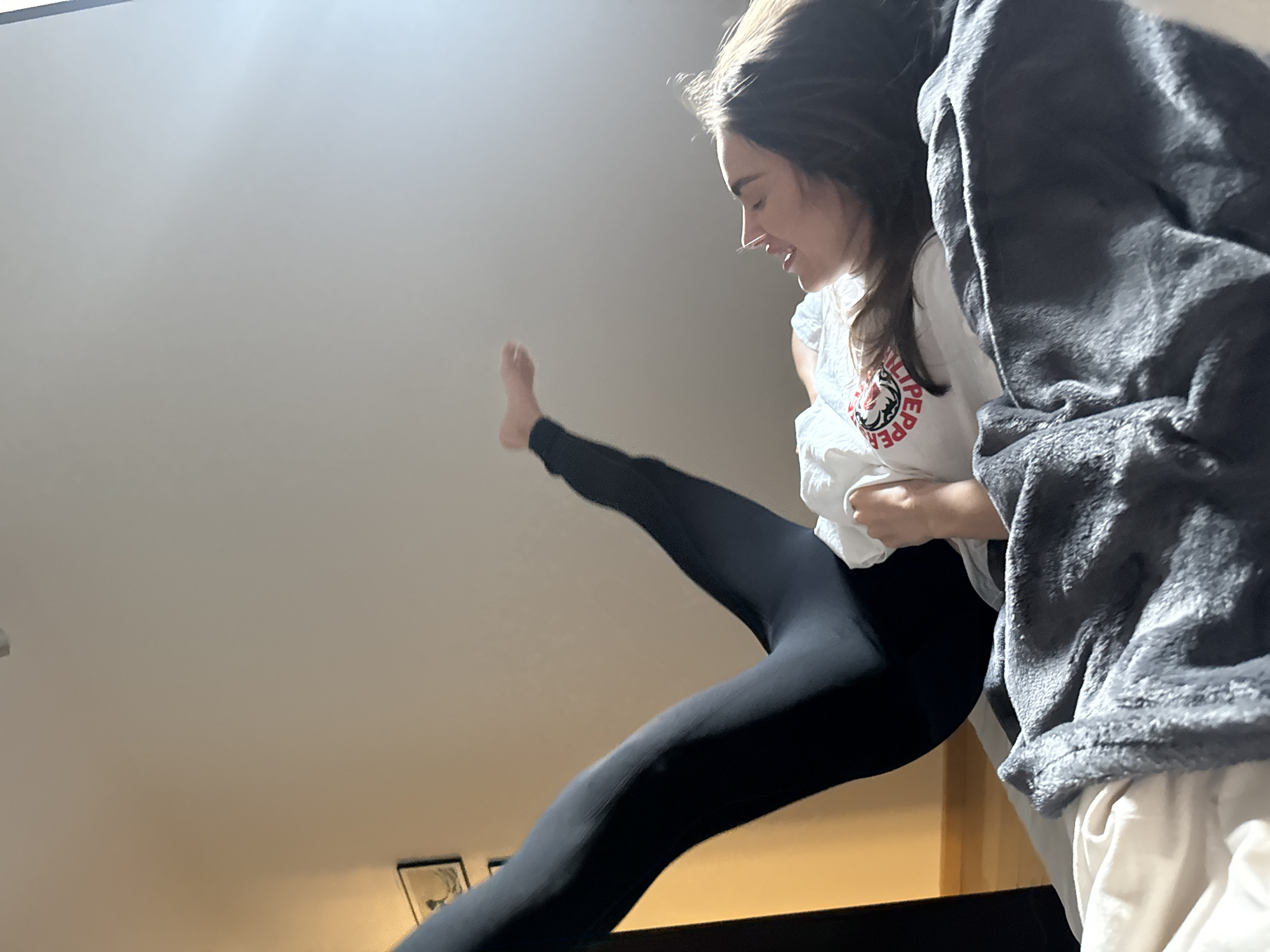 A woman in black leggings and a white t-shirt is executing a high kick indoors. She is smiling and seems to be in a playful mood. The room has a light-colored ceiling and walls, with some framed pictures visible in the background. A grey blanket is draped near her. The location is Unknown, Unknown.