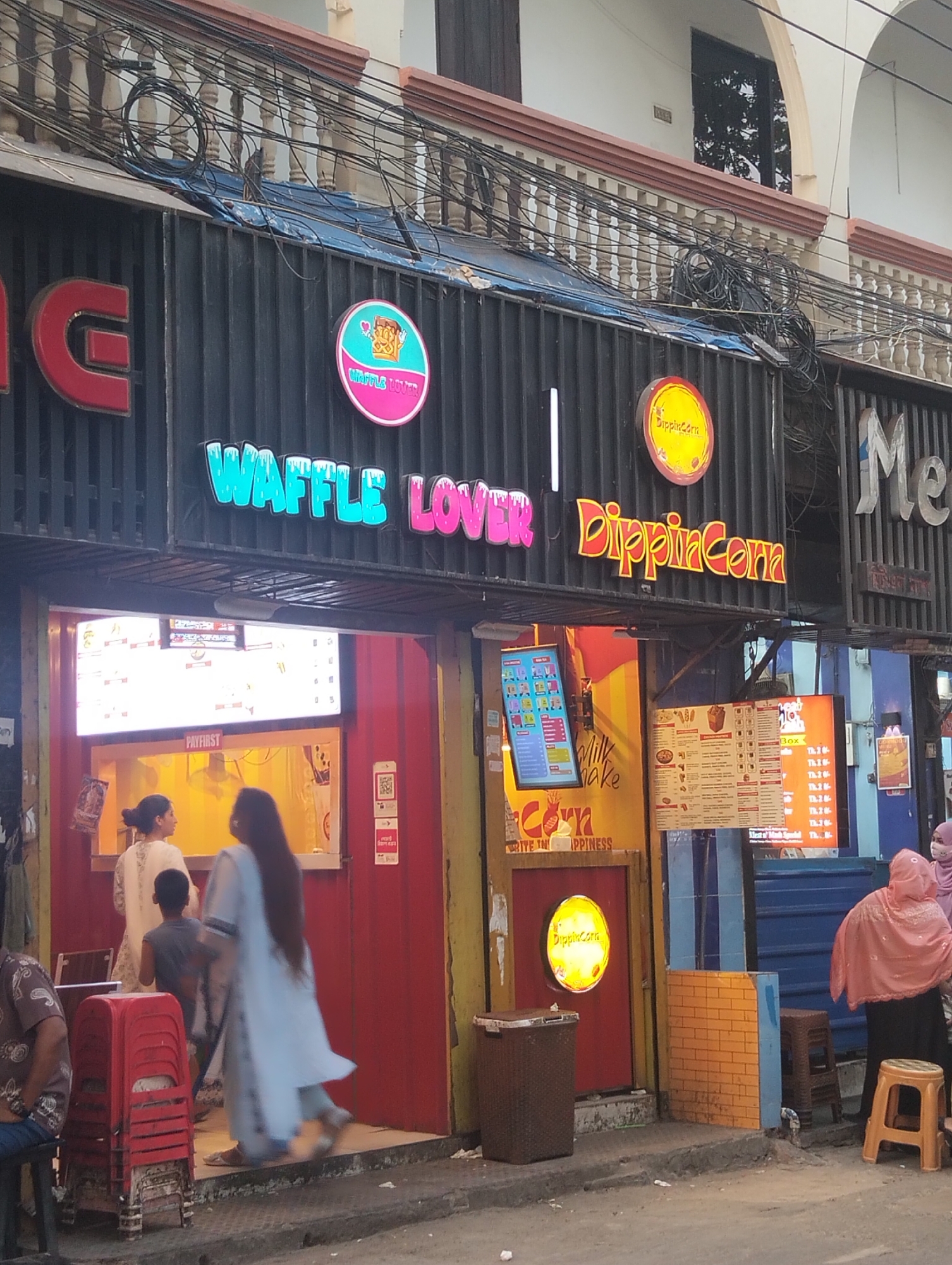 The image shows the storefront of "Waffle Lover | Dippin Corn" in Dhaka, Bangladesh. The store is located on a street with overhead electrical wires visible. The store front features signage in a modern font with neon colors on a black corrugated background. A couple and child are visible inside the establishment. A woman in a pink head covering and other clothing is outside of the store. Stools are stacked near the left entrance. Near the right entrance, there is a trash can and more stools. Menu boards are visible inside the storefront.