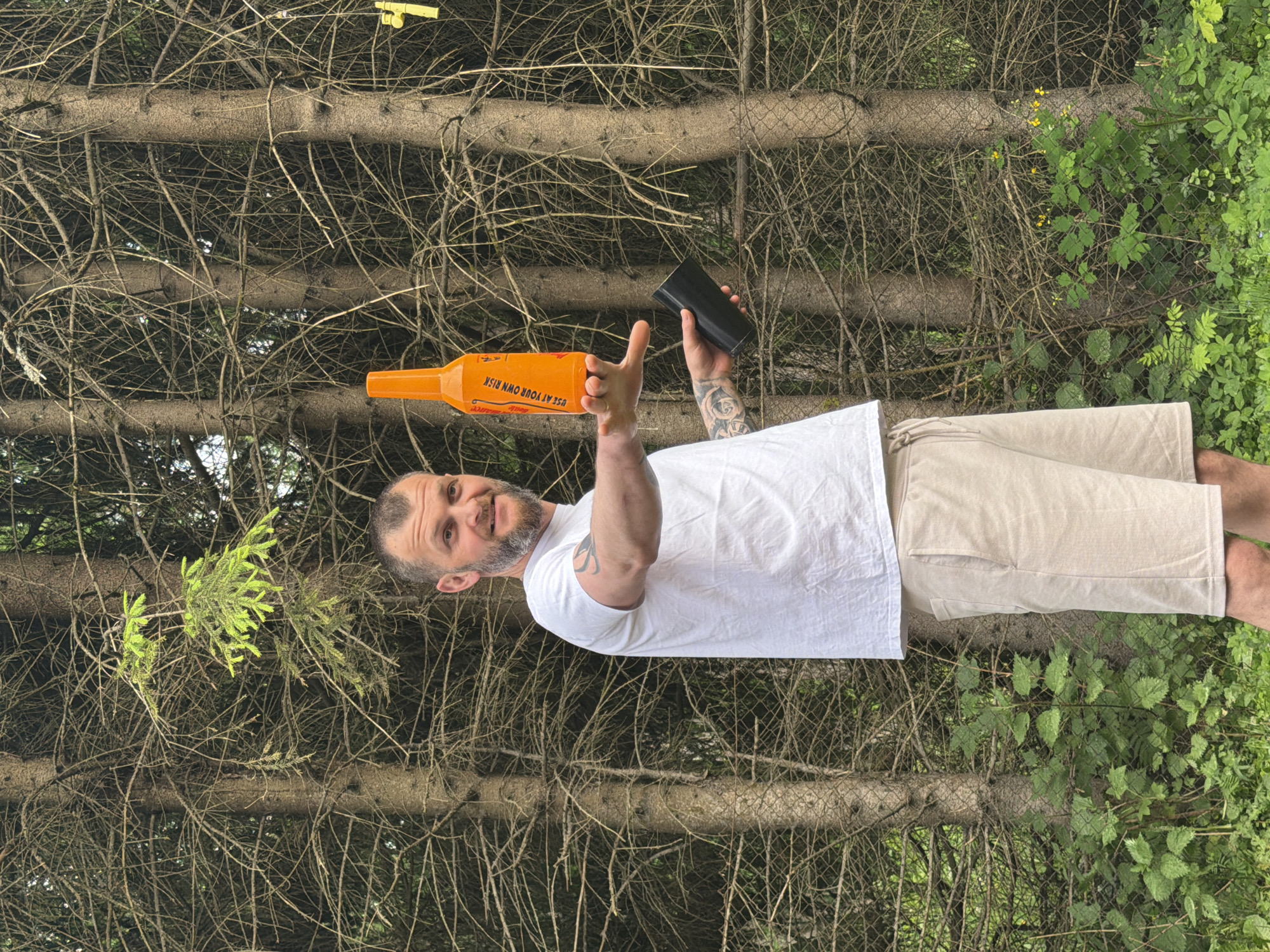 A Caucasian male with short brown hair and a beard, wearing a white t-shirt and light-colored shorts, stands outdoors in front of a dense forest of pine trees. He is holding an orange bottle in his right hand, with text visible on the bottle, and a black cylindrical object in his left hand. The man is looking towards the camera with a slight smile. The background consists of the thick trunks and branches of pine trees, with some green foliage and a wire fence visible at the bottom. The scene appears to be in a natural, wooded area.