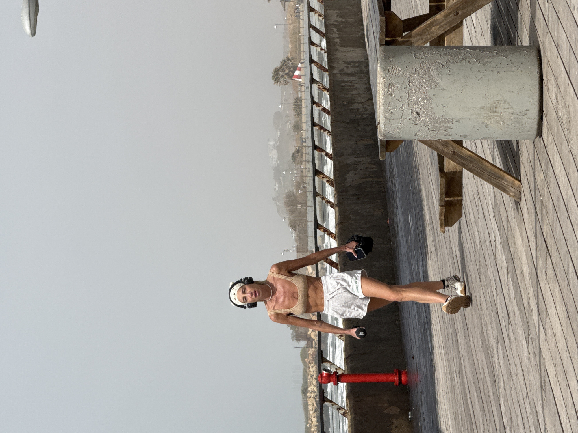 A woman exercises outdoors on a wooden deck. She wears a sports bra and shorts and uses dumbbells. The background shows the city of Tel-Aviv, Israel, with a coastline visible in the distance. A red fire hydrant is visible near the deck.