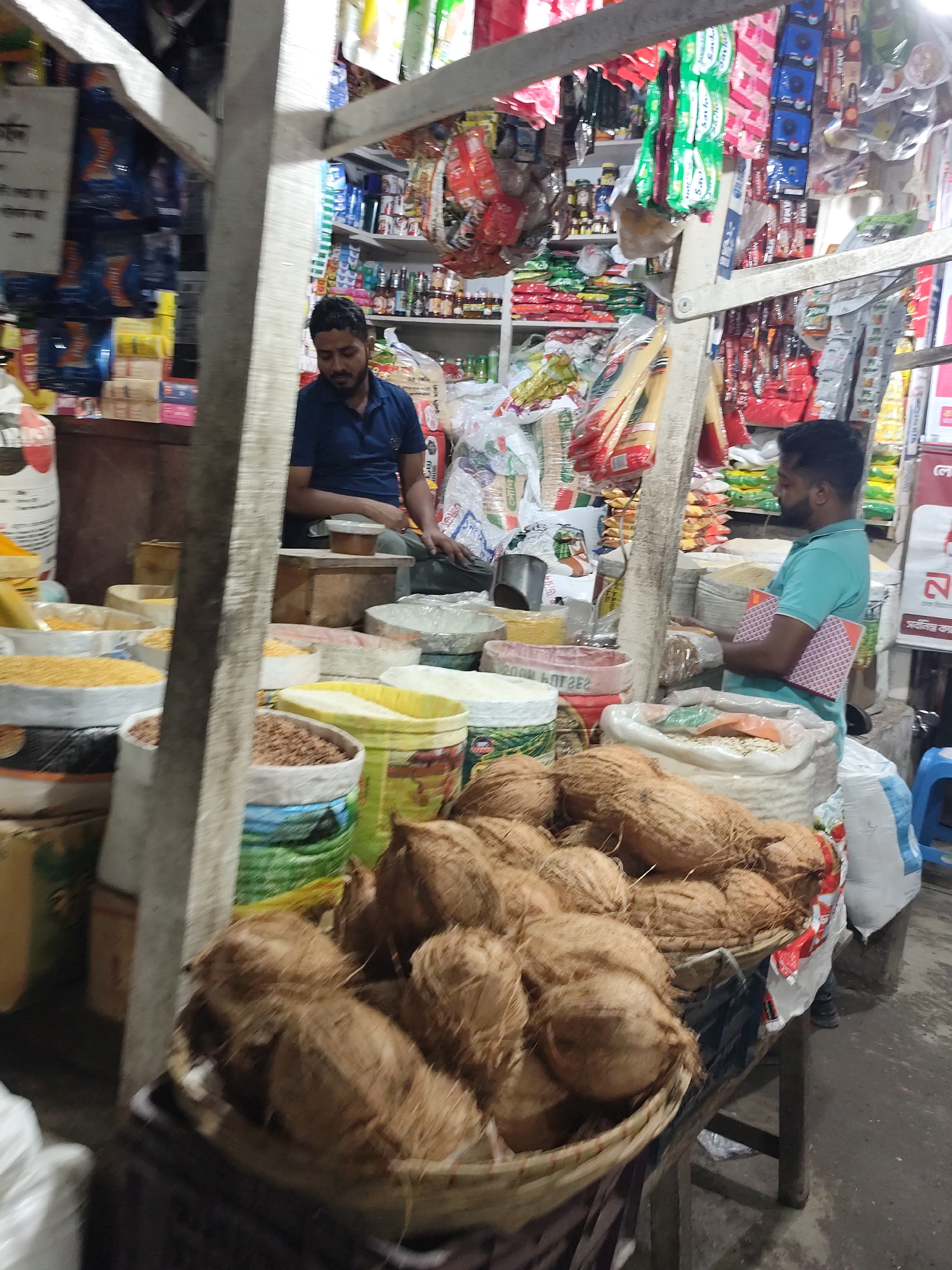 The image depicts a market scene in Dhaka, Bangladesh. Two South Asian men are present. The man on the left is sitting and wearing a navy blue short-sleeved shirt. The man on the right is standing and wearing a light turquoise shirt, holding a pink and red notebook. In the foreground, baskets are filled with coconuts. Other goods such as grains, flours, spices, and various packaged foods are visible on shelves and in sacks behind the men. The market appears to be covered with a makeshift roof supported by wooden beams.