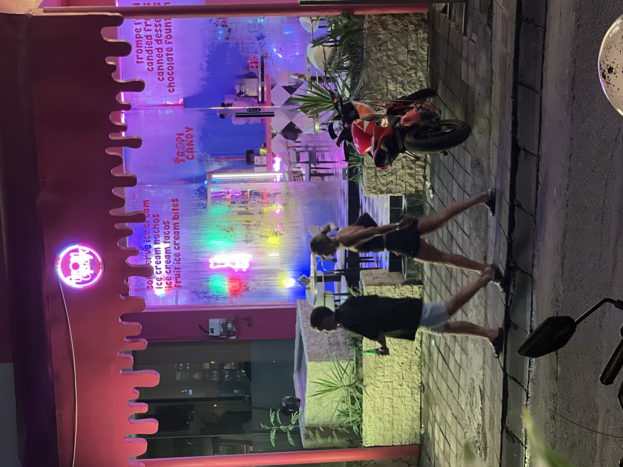 This night-time image captures a lively street scene in North Kuta, Indonesia, dominated by a brightly lit dessert shop named "TROPI CANDY." The shop's facade features a distinctive pink, "melting" or "dripping" design, adorned with a circular neon sign displaying its name. Inside, visible through expansive glass windows, vibrant multi-colored lights illuminate the interior.
Menu items are displayed on the shop windows, offering a range of sweet treats including "soft serve ice cream," "ice cream nachos," "ice cream tacos," "fruit ice cream bites," "candied fruit," "canned desserts," and a "chocolate fountain."
On the paved sidewalk in front of the shop, two individuals are walking. The person closer to the camera wears a dark top and light shorts, holding what appears to be a green bottle or container. Another individual in dark clothing walks further down the sidewalk. A red and white Honda motorcycle is parked on the sidewalk, positioned between the colorful shop and the street. The sidewalk is constructed from light-colored stone blocks, and features planter boxes with green foliage. The dark asphalt of the road is visible on the right side of the frame. The scene conveys a casual, bustling evening atmosphere.