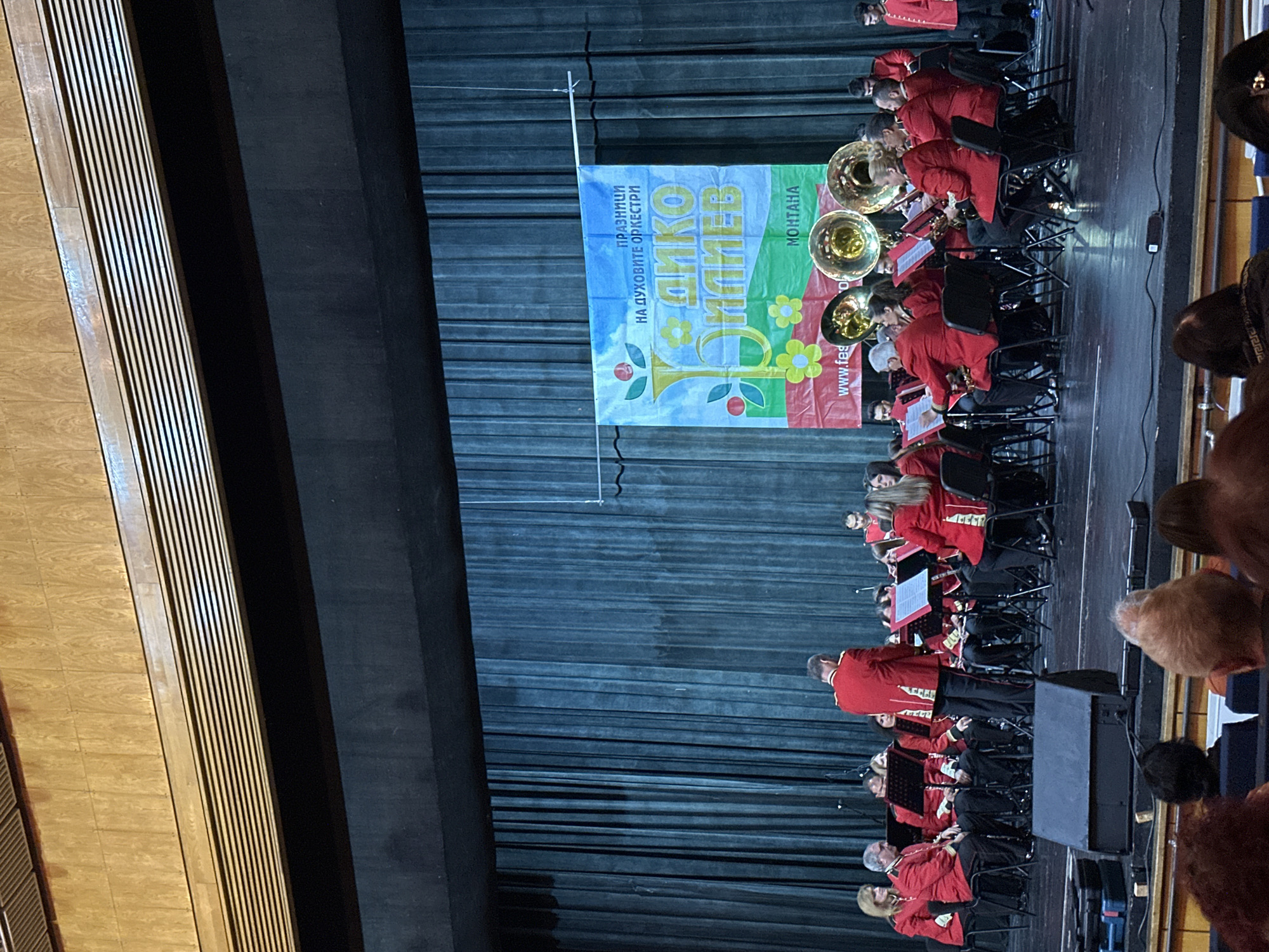 A stage in Montana, Bulgaria features a brass band in red uniforms performing in front of a banner. The banner is in Bulgarian and reads "Празници На духовите оркестри, ДИКОВ Монтана", which translates to "Festival of Brass Bands, Dikov Montana". Audience members are seated in front of the stage. The stage has wood paneling on one side and a dark curtain backdrop.