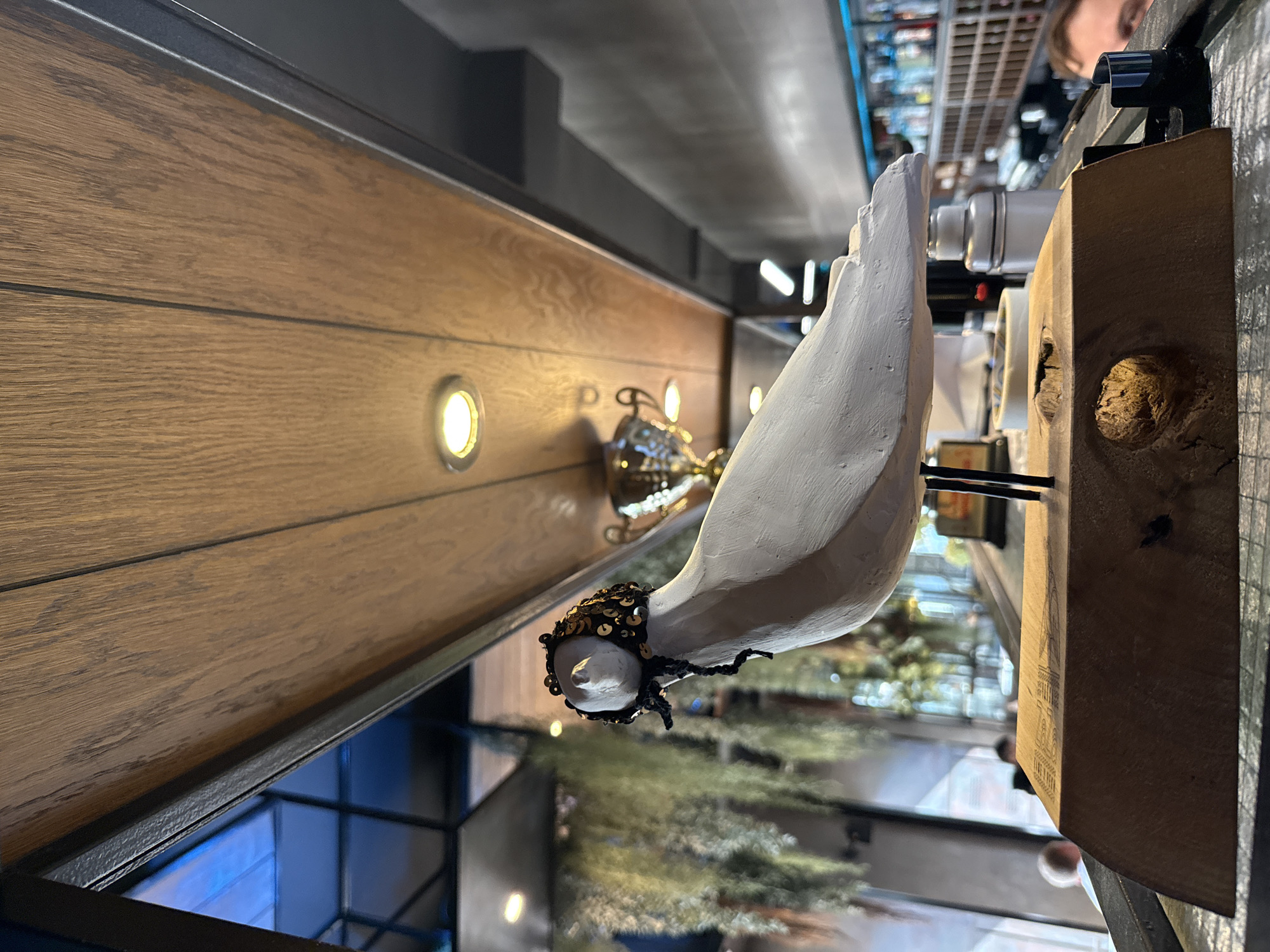 This is a close-up, low-angle shot of a white, bird-shaped sculpture with a black, sequined cap adorned with gold sequins and black ties. The sculpture is mounted on a piece of wood with an irregular hole. The background is blurred, showing a ceiling with wooden panels and circular lights, a shelf with hanging plants, and a bar area with bottles and a shelf. The image does not provide any information about the city of Perm, Russia.