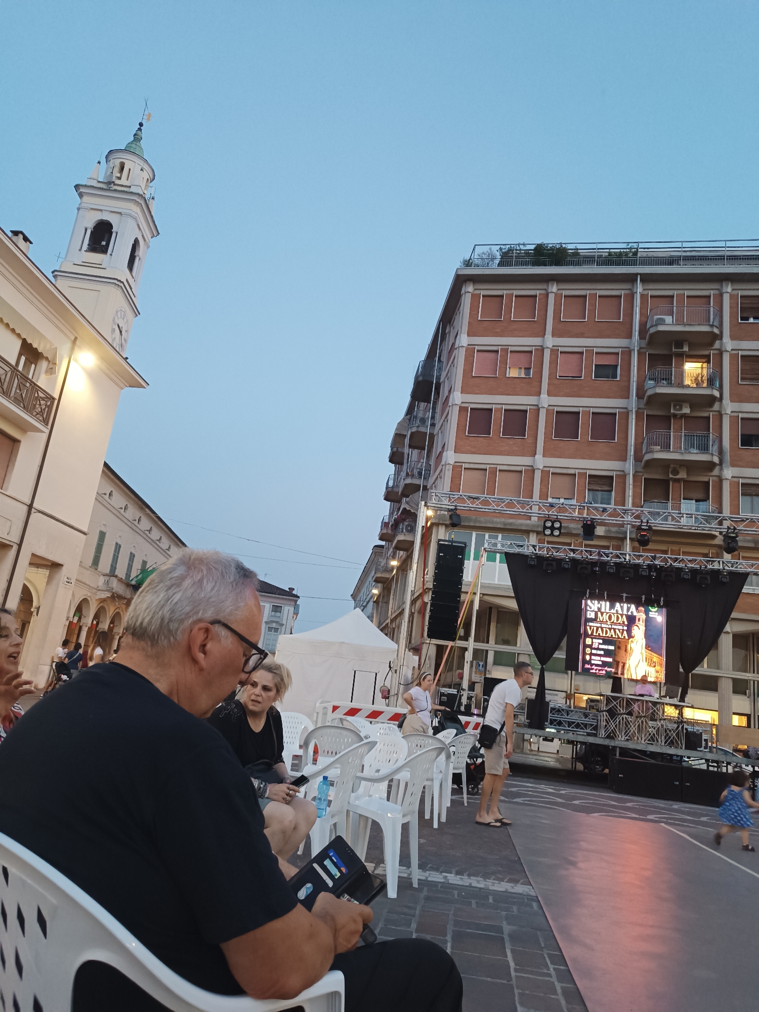 In Viadana, Italy, an outdoor event is underway. The sky is a clear twilight blue. In the background, a multi-story brick building with balconies and numerous windows dominates the skyline. In front of it, a stage is set up with a large screen displaying "SFILATA DI MODA VIADANA" along with text in Italian and an image of a woman. Lights are positioned above the stage.
To the left of the stage, a white tent is visible. Several white plastic chairs are arranged in front of the stage, with some occupied by people. In the foreground, a man with glasses and graying hair is seated, looking at a tablet. To his right, a woman with blonde hair is seated, also appearing to be engaged with her phone.
Further to the left, a white bell tower with a clock face rises against the sky, a characteristic architectural feature of Italian towns. The street is paved with cobblestones, and there are awnings and windows on the buildings lining the street. A young child in a blue dress is running on a grey surface in front of the stage. The overall atmosphere suggests a public gathering or festival.