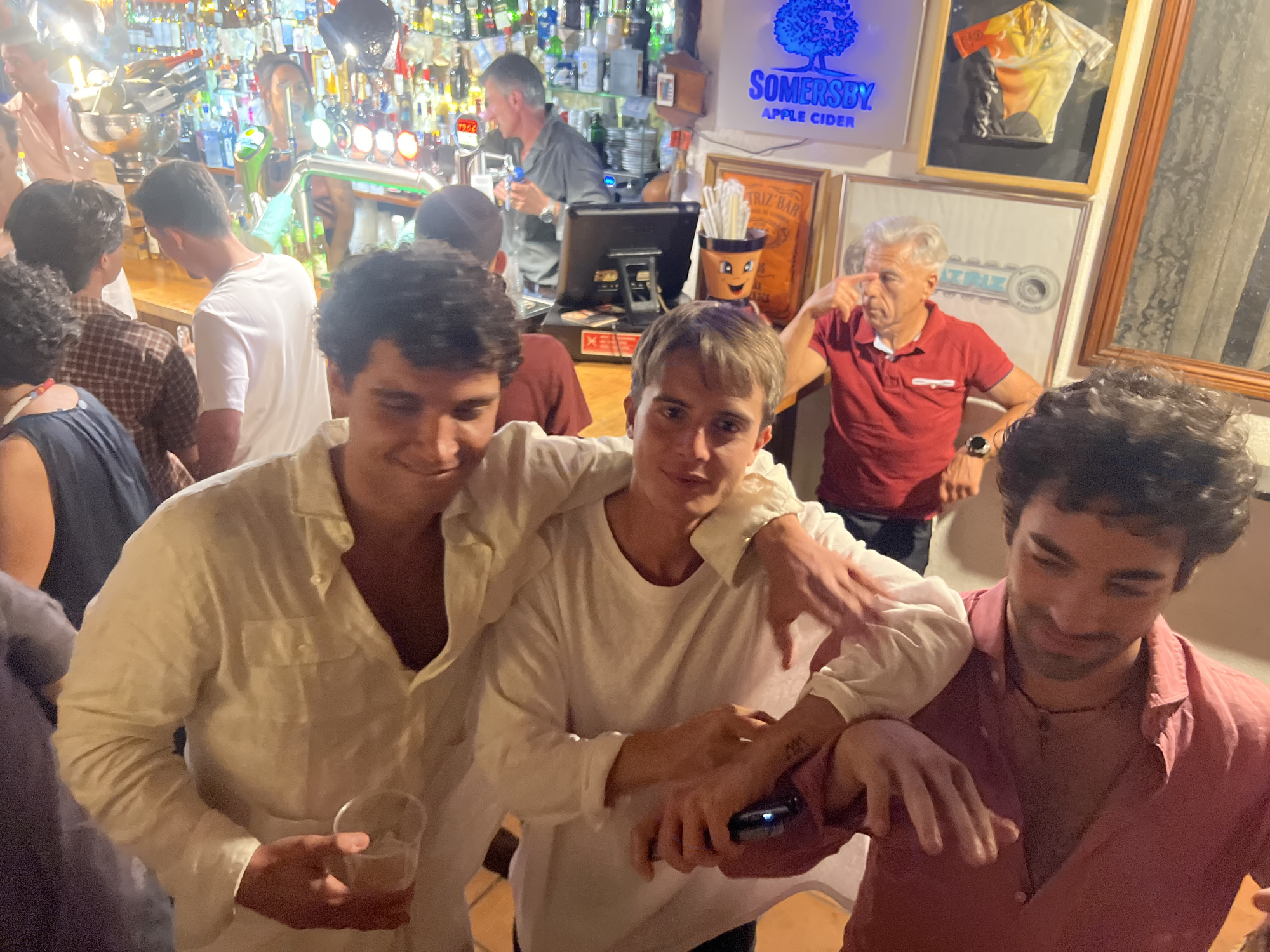 The image captures a lively indoor gathering, likely a bar or restaurant, with a well-stocked bar in the background featuring numerous bottles of alcohol and ambient lighting. In the foreground, three young men are the primary subjects. Two of them, positioned in the center and to the left, are close together, with their arms around each other, suggesting camaraderie. Both are wearing light-colored, loose-fitting shirts. The man on the left is holding a clear plastic cup with a beverage. The man on the right is reaching out with his left hand, touching the arm of the man beside him, and appears to be holding a smartphone in his right hand. A third young man, on the right side of the frame, is wearing a pink collared shirt and is looking downwards, with his left arm resting on the shoulder of the man in the pink shirt. An older man with grey hair, wearing a red polo shirt, is visible in the mid-ground, pointing with his finger. The environment appears to be bustling with activity, indicated by the presence of multiple people and the implied social interaction. The presence of a "SOMERSBY APPLE CIDER" sign on the wall and the overall atmosphere suggest a casual and celebratory occasion. While the specific location in Caminha, Portugal is not visually confirmed within the image itself, the context implies a social establishment within that city.