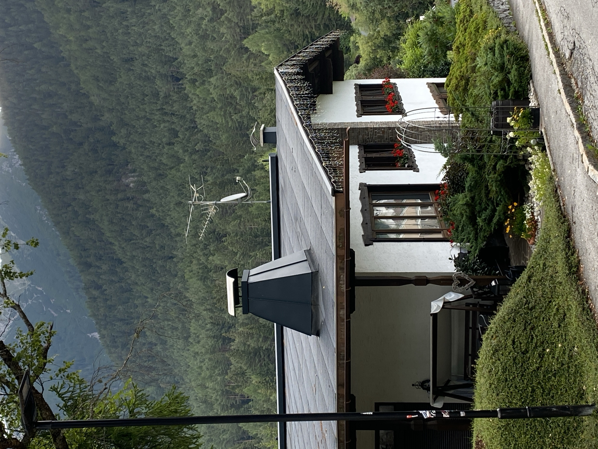 The image shows a house with a dark grey, sloped roof in Reith bei Seefeld, Austria. A large, dark grey pyramidal chimney is prominent on the roof. The house has white walls, dark wooden trim around the windows, and windows with red flowers in the window boxes. Decorative lights hang from the eaves of the roof. A black lamppost stands in front of the house, partially obscuring a porch with a swing set and a table covered with a white cloth. Lush green bushes and a small garden with yellow and white flowers are in front of the house. In the background, a dense, dark green forest covers a steep mountain slope. A satellite dish and an antenna are visible on the roof. The scene appears to be a residential property in a mountainous, forested area.