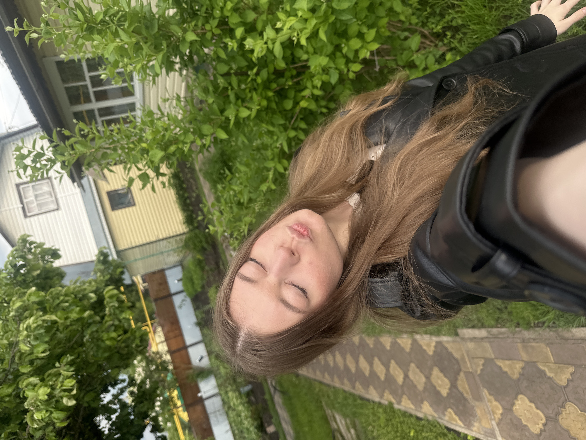 A young woman with long brown hair and closed eyes is posing for a selfie outdoors. She is wearing a black leather jacket. Behind her are green trees and parts of buildings. The ground appears to be a paved walkway with grass growing between the stones. The photo seems to be taken in Коноваловка, Russia, based on the location mentioned.
