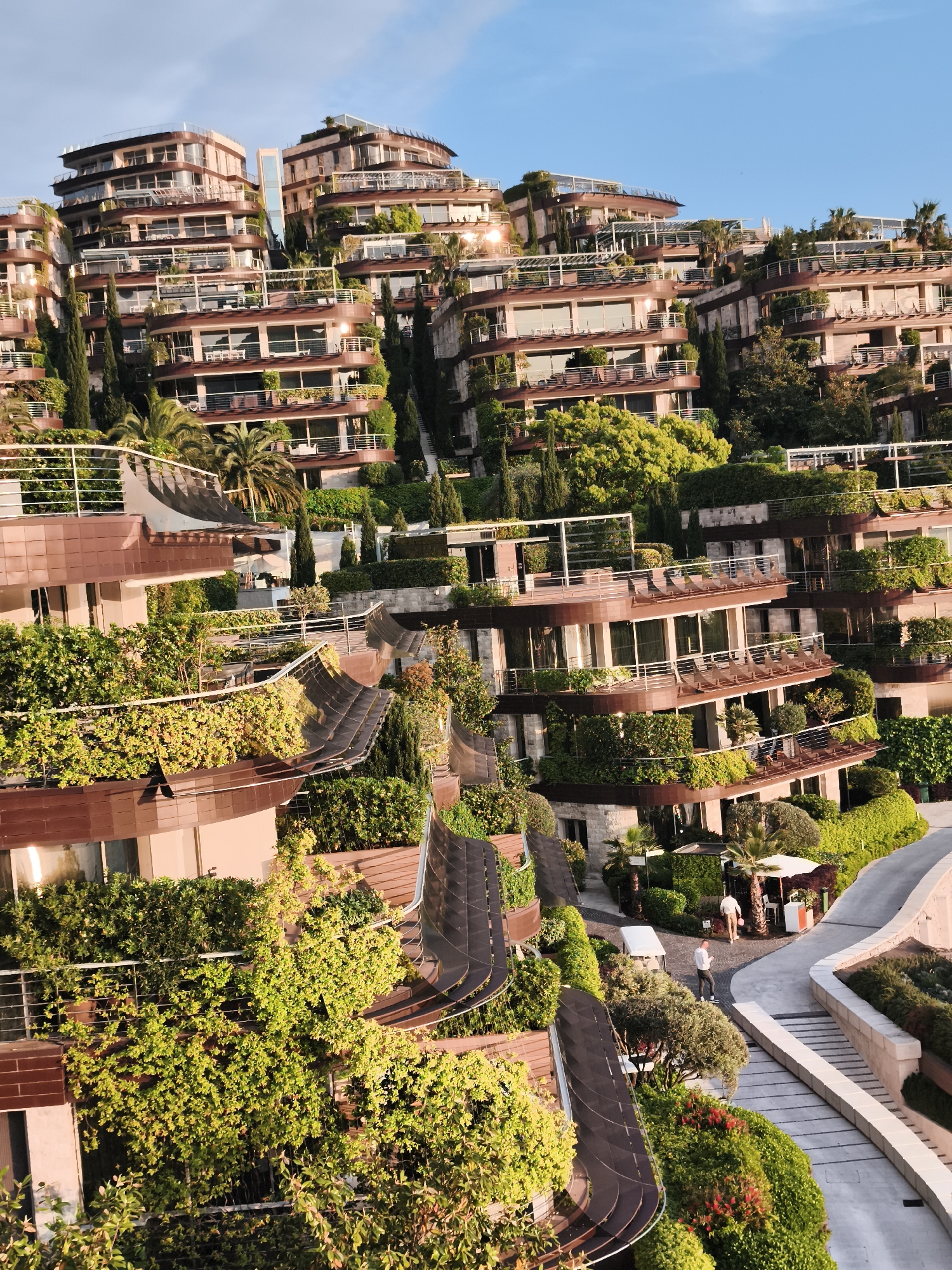 This image depicts a stunning terraced resort or residential complex nestled into a verdant hillside, characteristic of a Mediterranean landscape like Budva, Montenegro. The scene is dominated by multi-story buildings with a modern architectural style, featuring light-colored and brown facades, extensive glass windows, and numerous balconies with sleek glass railings.
A defining feature is the extraordinary integration of lush greenery with the architecture. Each level and balcony is extensively landscaped with a variety of trees, shrubs, and climbing plants, creating green walls and overflowing planters that blend the structures into the natural environment. Tall cypress-like trees and palms are interspersed throughout the foliage, adding to the picturesque setting. Winding pathways and stone steps connect the different levels of the complex. A prominent glass elevator shaft can be seen ascending between two sections of the buildings in the upper-middle.
In the lower right portion of the image, several people are visible on the pathways. One person in a white shirt and dark trousers stands near a small white golf cart, while two others are further back, near a white parasol, suggesting a leisurely environment.
The time of day appears to be either late afternoon or early morning, as indicated by the warm, directional sunlight that casts long shadows and brilliantly illuminates the building facades and vibrant greenery. The sky above is clear and blue. The overall atmosphere conveys luxury, tranquility, and a close connection to nature. No discernible text is visible within the image.