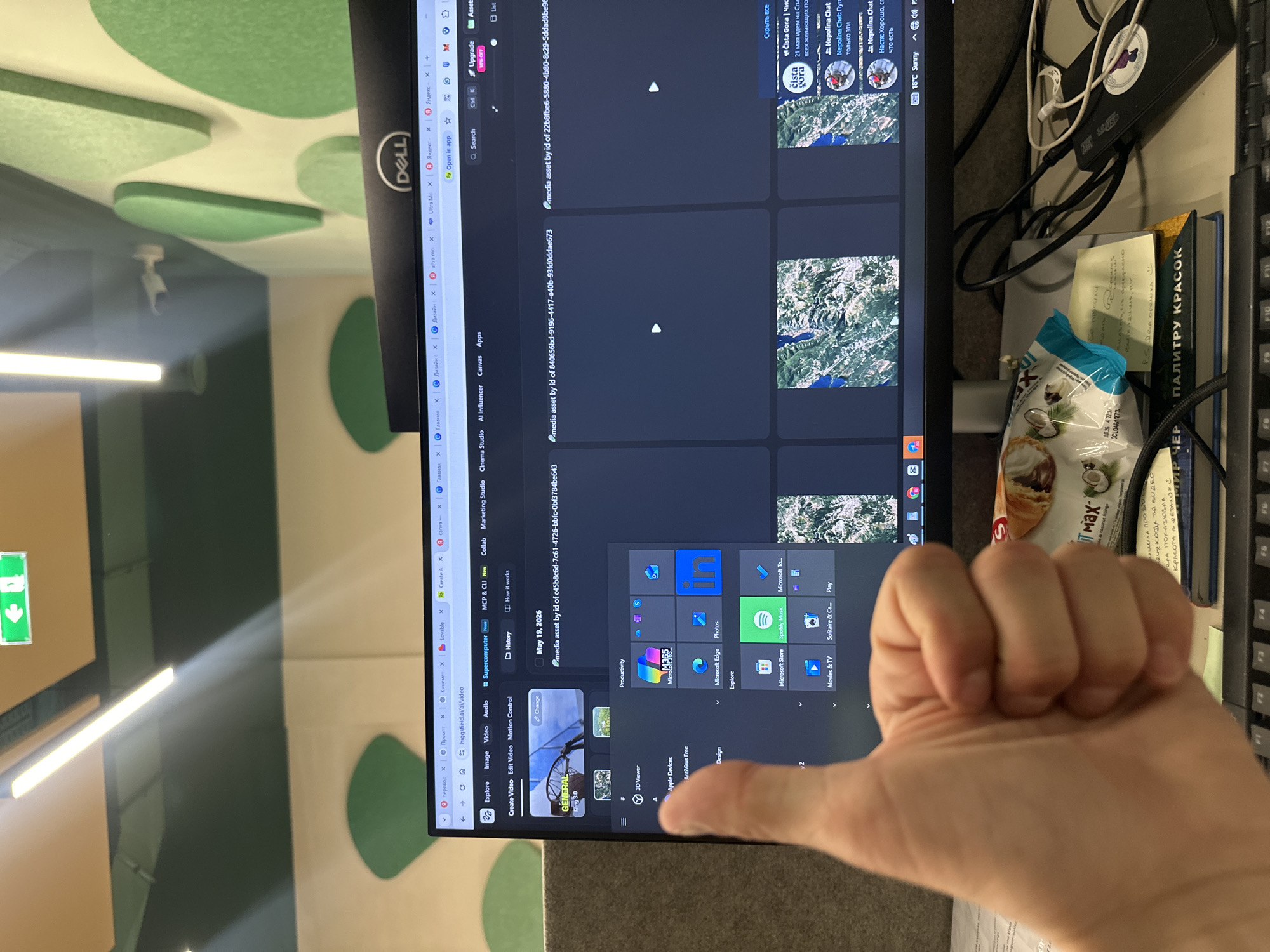 A person's hand is in the foreground. Behind the hand, there is a Dell computer monitor. The monitor displays the Microsoft Windows operating system, with various applications and web pages open. A book titled "ПАЛИТРУ КРАСОК" and a packet of "MAX" coconut fillings are visible to the right of the monitor. In the background, there is a wall with green soundproofing panels of various shapes. There is a light fixture attached to the ceiling. The text on the screen suggests the user is accessing or working with media assets and related applications. The images on the screen of "Cista Gora", along with Cyrillic text referencing Napolina Chat, indicate a location in Montenegro. The presence of the city of Budva cannot be confirmed.