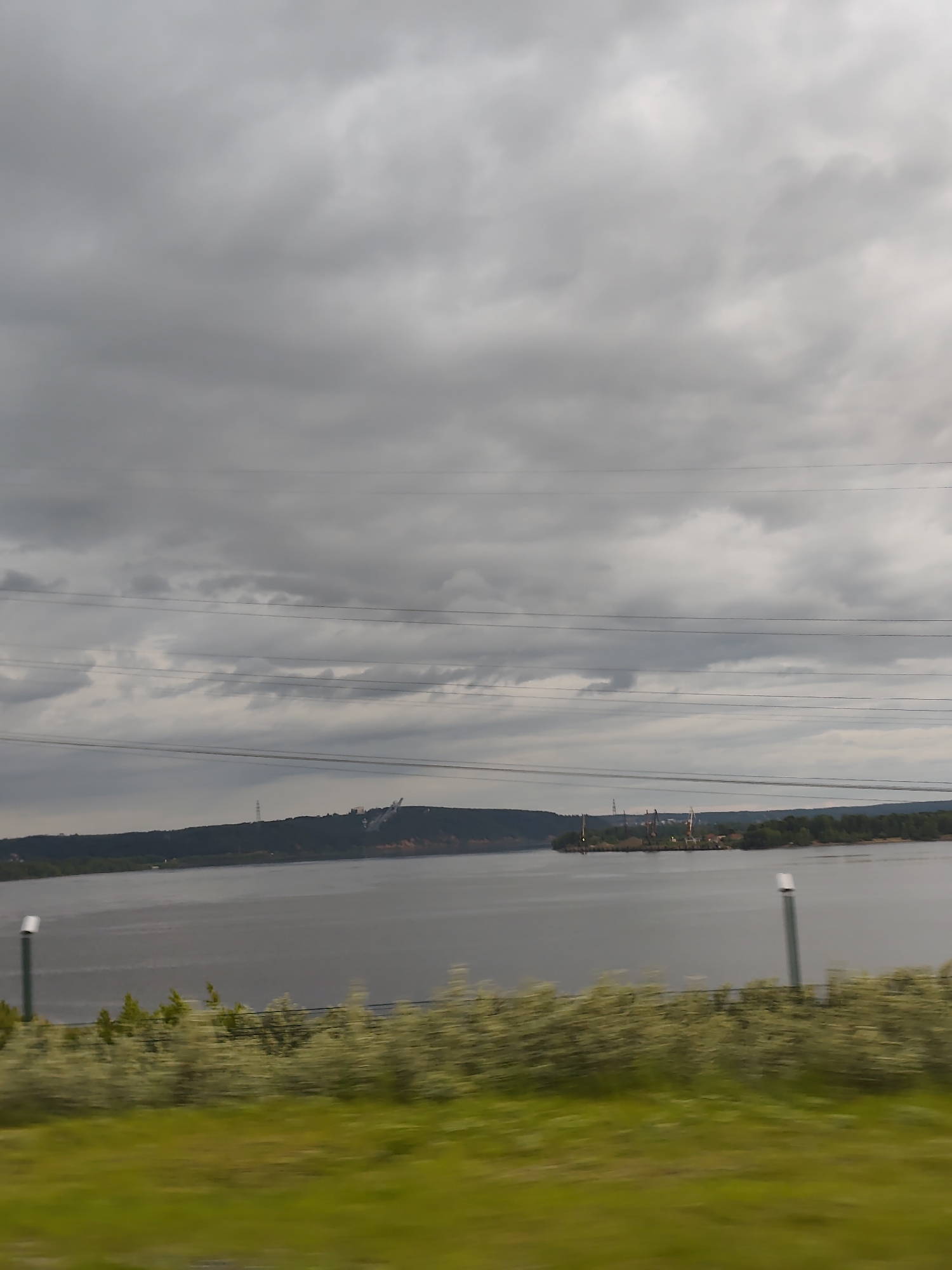 This image captures an overcast, industrial and natural landscape scene in the city of Chaikovsky, Russia, likely viewed from a moving vehicle. The sky occupies approximately the upper two-thirds of the frame, filled with dense, uniform grey clouds indicative of an overcast day. Several dark, parallel power lines or communication cables traverse horizontally across the upper midground, partially obscuring the distant landscape.
The foreground, approximately the bottom fifth of the image, features bright green grass and blurred light green or white-leafed bushes, heavily streaked horizontally due to motion blur. Two vertical poles, one dark green with a white top on the left and another similar one further right, are partially visible in the blurred foreground.
The midground is dominated by a wide, calm body of water, likely a river or lake, reflecting the grey sky. Across the water, the distant shorelines are visible. On the left background, a forested landmass or hill rises, upon which a tall, slender lattice tower stands. To the right of this tower, a large, prominent white and grey structure, resembling a ski jump or an industrial ramp, ascends the hillside.
On the right background, another forested landmass extends into the water, forming a small peninsula or island. On this landmass, a cluster of industrial structures is visible, including multiple cranes (some red-tipped), various buildings, and what appears to be a port or a factory complex situated directly at the water's edge. No people, specific actions, or interactions are discernible within the frame.