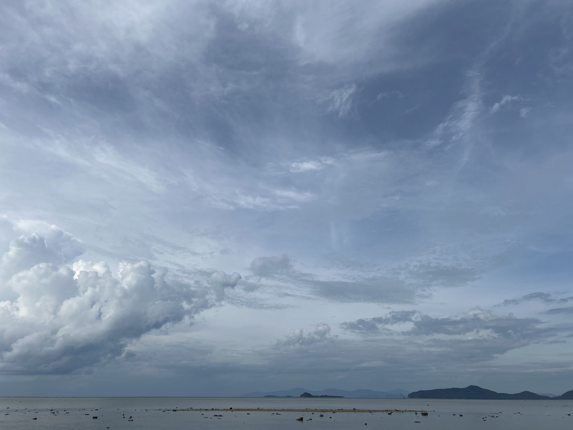 The image depicts a seascape in Baan Harn, Thailand, under a sky filled with varying cloud formations. To the left, large cumulonimbus clouds loom, while the rest of the sky is covered in cirrus and altostratus clouds. The water's surface is relatively calm, with small rocks and land partially submerged. In the distance, islands are visible on the horizon. The overall light suggests an overcast day. No people are visible in the frame.