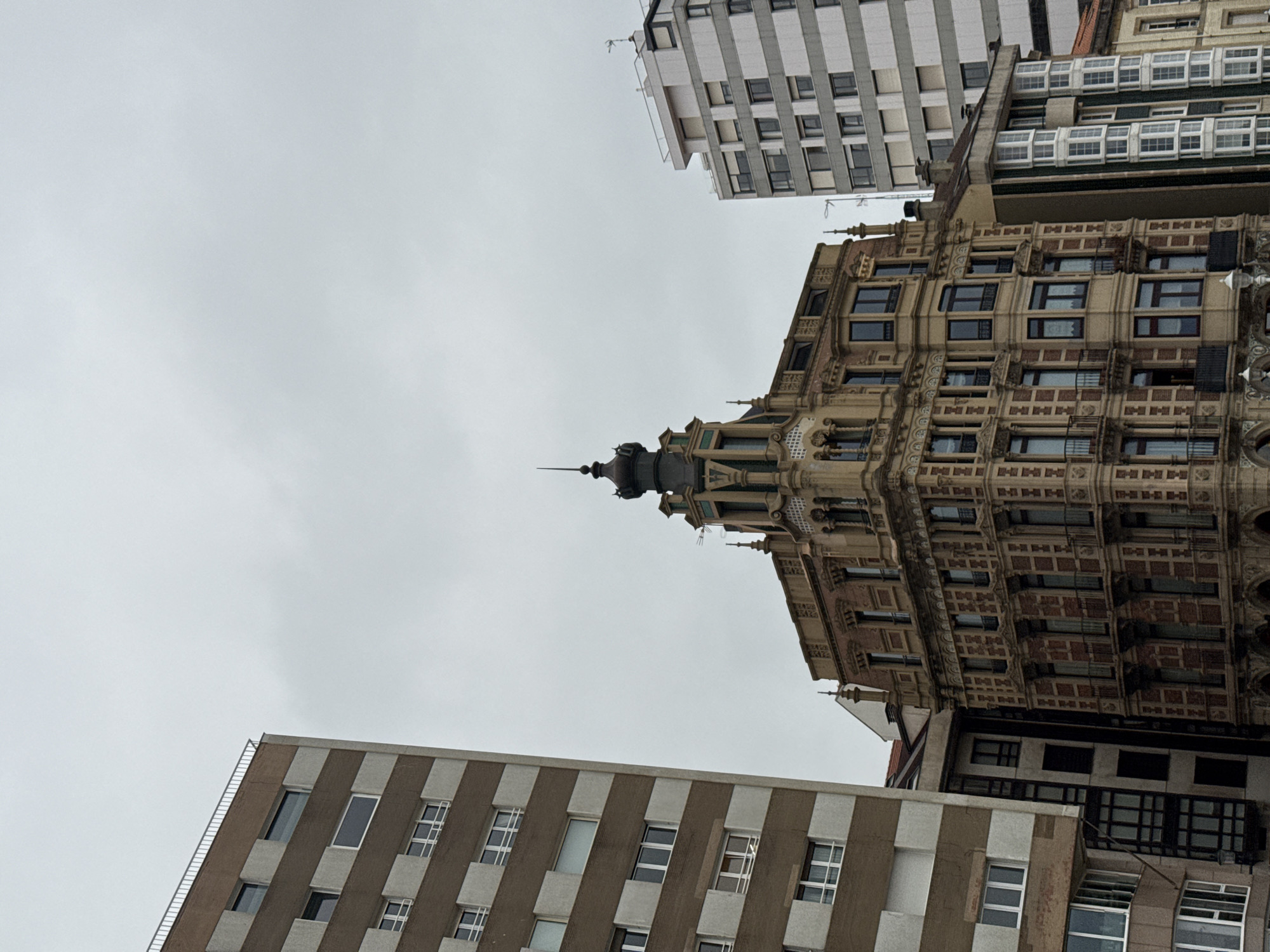 The image shows a low-angle view of buildings in Gijón/Xixón, Spain against an overcast sky. The main building is ornately decorated with a tower and pointed spire. Surrounding buildings include taller modern structures with repetitive window patterns in either gray and white or brown and white. There are no people visible.