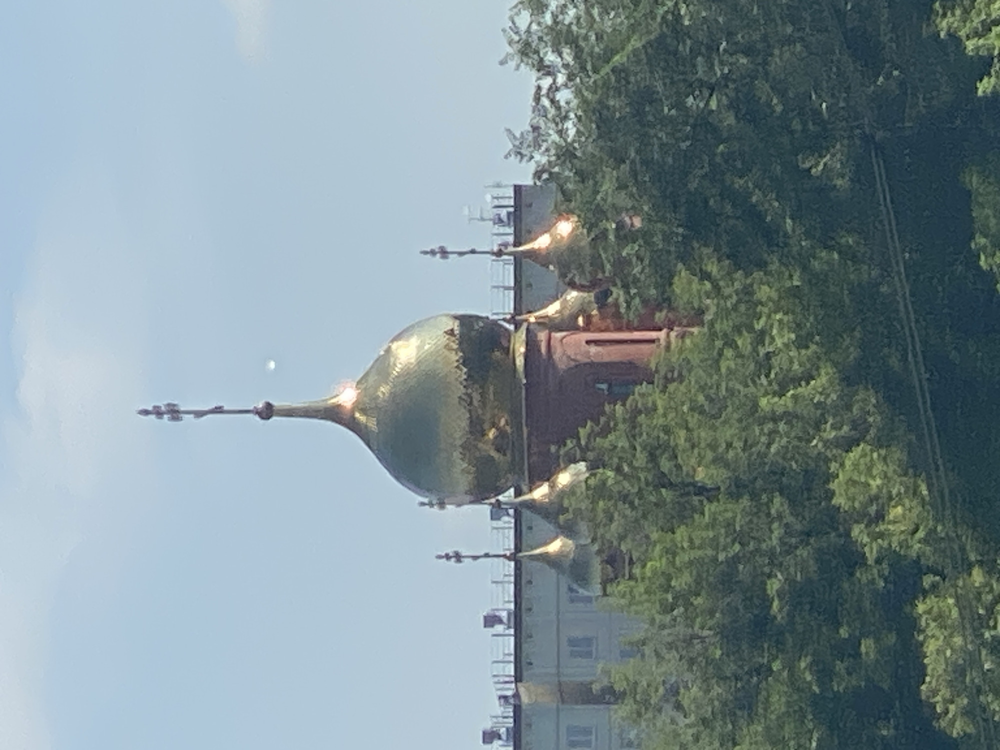 A bright, sunny day in Novokuznetsk, Russia, features a close-up, eye-level, medium shot of a red brick church adorned with multiple golden domes, each topped with a cross. The largest dome is in the center, with smaller domes to its left and right. Behind the church, a modern building with windows and a flat roof is visible, partially obscured by lush green trees. The sky is clear blue with a few wispy white clouds and a faint luminous object, possibly a distant aircraft or celestial body, can be seen in the upper right quadrant of the frame. The focus is on the church's distinctive architecture and its interplay with the surrounding urban and natural elements.