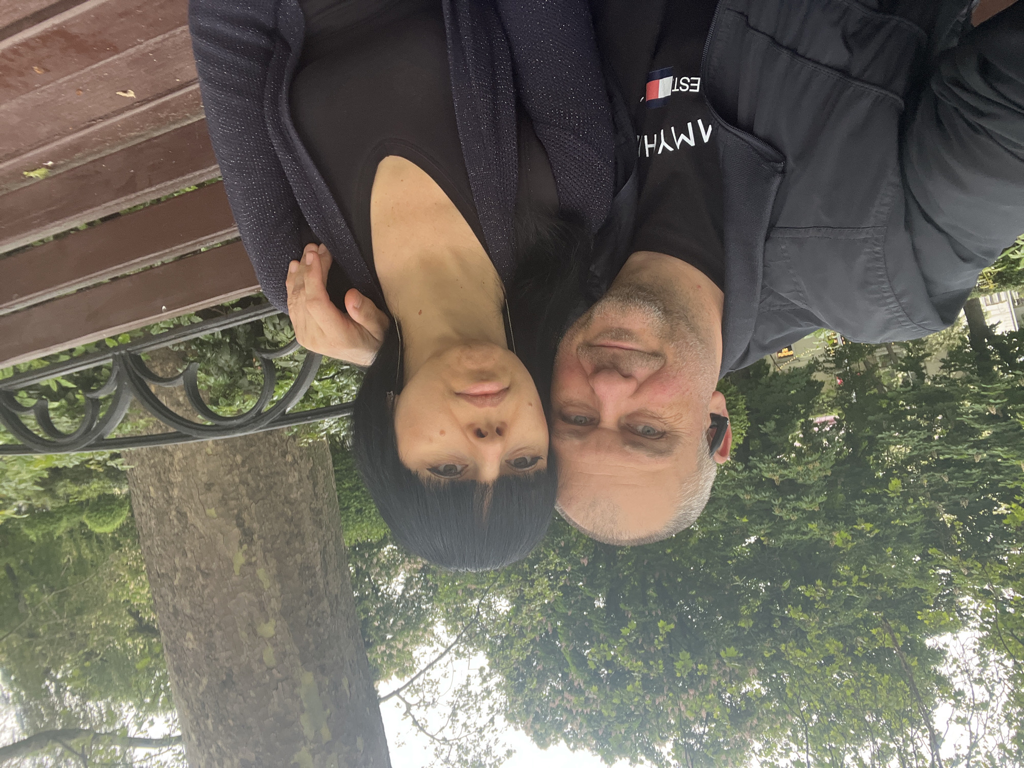 A couple is photographed close together on a bench in what appears to be a park in Krasnodar, Russia. The woman has dark hair and is wearing a dark cardigan over a dark shirt. The man has short gray hair and is wearing a black t-shirt with the Tommy Hilfiger logo and a black jacket. They are positioned shoulder to shoulder, looking directly at the camera. The background includes trees and foliage suggesting a park setting. The bench appears to be made of wood with a metal frame.