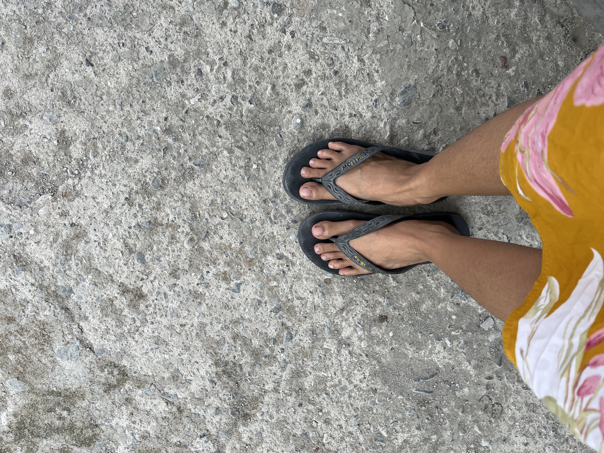 A first-person perspective shot shows a person's bare feet in dark gray flip-flops standing on a rough, textured gray concrete surface. The concrete is uneven with visible aggregate and some cracks. The person's legs and a portion of their colorful shorts, featuring a floral pattern with pink and yellow elements on an orange background, are visible in the lower part of the frame. The scene is outdoors with no specific identifiable landmarks to confirm the location in Kemangguhan, Indonesia.
