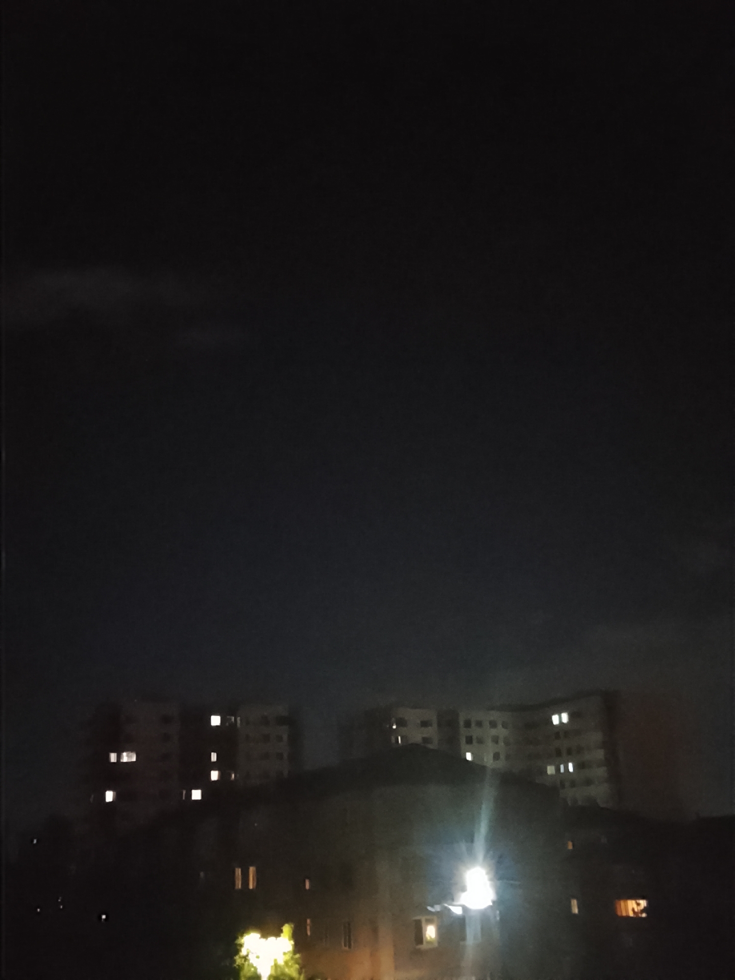 The media file presents a dark, nighttime scene of a residential area in Yerevan, Armenia. The sky occupies the upper two-thirds of the frame, appearing very dark with subtle variations in tone, suggesting either a clear night with some atmospheric haze or faint, high-altitude cloud cover.
In the middle and background, several large, multi-story buildings, consistent with apartment blocks, are visible. Their structures are mostly silhouetted against the dark sky, but numerous windows emit bright, artificial light, appearing as glowing rectangles and squares. On the left side, one building displays several lit windows, while a larger building on the right also shows multiple illuminated units.
The foreground and lower mid-ground are characterized by dark, indistinct shapes that suggest trees or dense foliage, partially obscuring the foundations and lower floors of some buildings. A prominent, intensely bright, and somewhat diffused light source is visible in the lower right, possibly a streetlamp or a strong interior light, creating a significant lens flare effect. Another smaller, yellowish light is discernible in the lower left.
No individuals or specific activities are visible within the frame. The scene conveys a sense of quiet urban stillness typical of late night hours. There is no discernible text in the image. The architecture of the visible buildings aligns with the provided location context of Yerevan.