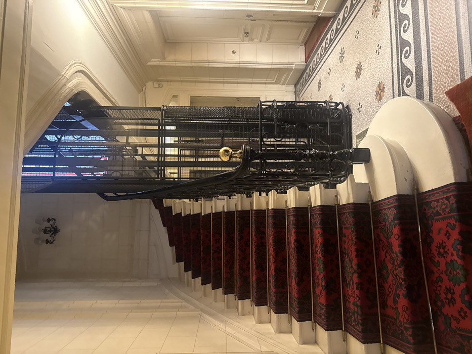 An interior view depicts a grand entrance hall within a historic Parisian building, characteristic of late 19th or early 20th-century Haussmannian architecture. A wide, curving staircase, constructed of white marble or stone, ascends to the left, adorned with a dark red patterned carpet runner secured by brass rods at each tread. To the immediate right of the staircase, a vintage, open-cage elevator shaft crafted from dark wrought iron features a delicate, multi-tiered design, culminating in a prominent brass finial at its base on the ground floor.
The ground floor is paved with an elaborate mosaic tile pattern, incorporating white, grey, black, and muted earth tones in geometric and floral motifs. The walls are paneled and painted in an off-white or cream color, featuring classic decorative moldings. Two distinct white paneled doors, each with a brass knob, are visible on the far right, set within the wall. Overhead, a dark metal, multi-bulb wall sconce is affixed to the wall above the curving staircase. No individuals or active interactions are visible within the scene. The lighting suggests an interior space, potentially with ambient natural light filtering from an unseen source beyond the elevator cage.