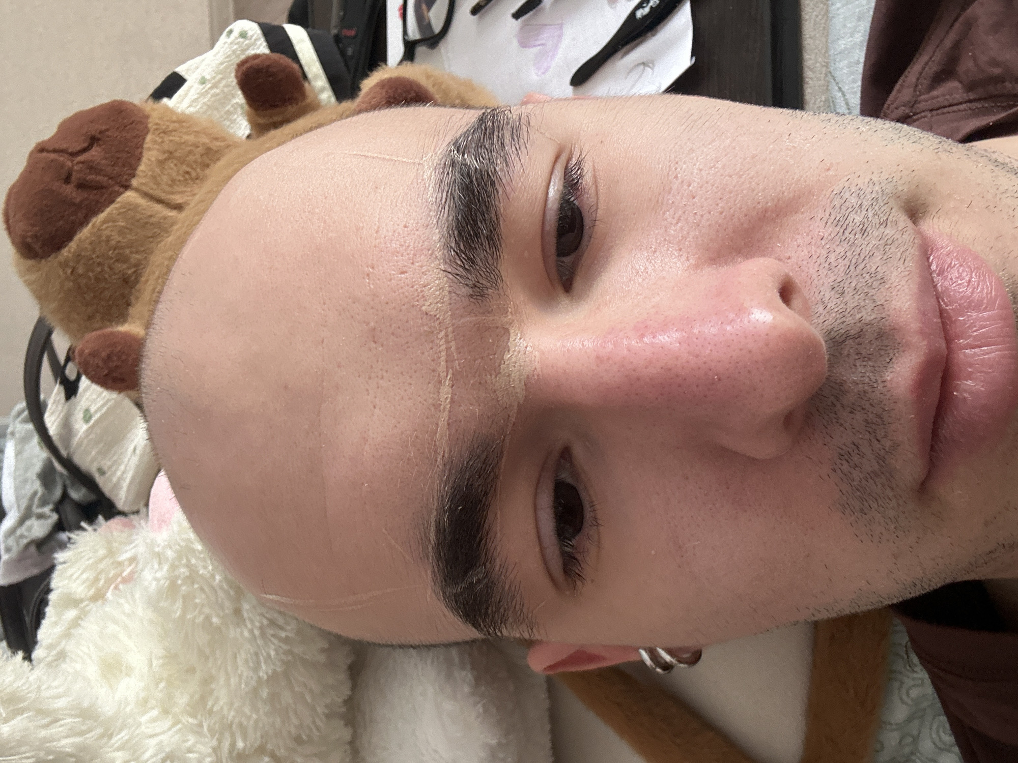 A close-up, horizontally-oriented shot shows a person's face lying on what appears to be a bed. The person has a shaved head with a hairline drawn on the forehead and thick eyebrows. The skin tone is light, and there is stubble on the cheeks and around the mouth. The person's eyes are open. The lower ear is visible, pierced with a silver hoop earring. A brown stuffed bear is positioned near the person's head. A white, fuzzy stuffed animal is nearby as well. Background elements include what appears to be bedding and other miscellaneous objects, possibly cosmetic items. There is no indication of the location being Aktanysh, Russia.