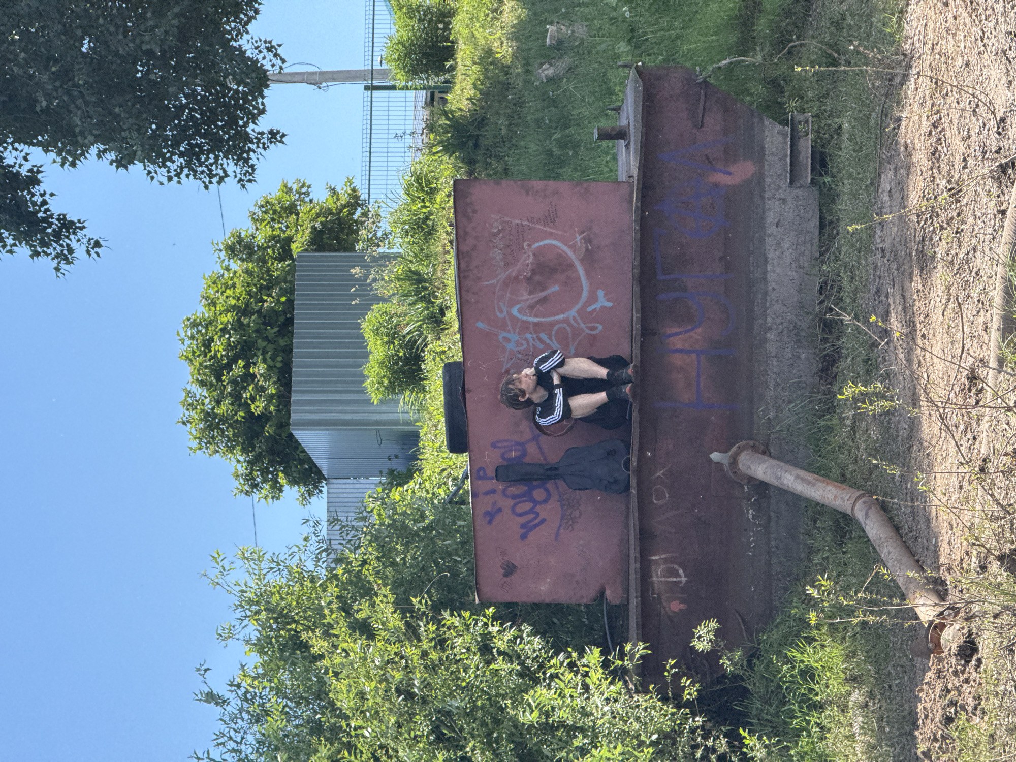 A young person with light brown hair sits on a large, red, metal structure covered in graffiti, next to a black guitar case. They are wearing a black t-shirt with white stripes on the sleeves and black shorts. The setting appears to be an outdoor industrial or abandoned area. Green trees and vegetation surround the structure, and a clear blue sky is visible above. In the background, there is a metal fence and what appears to be corrugated metal sheeting. A rusty pipe extends from the ground towards the metal structure. The location is identified as Chaikovsky, Russia.
