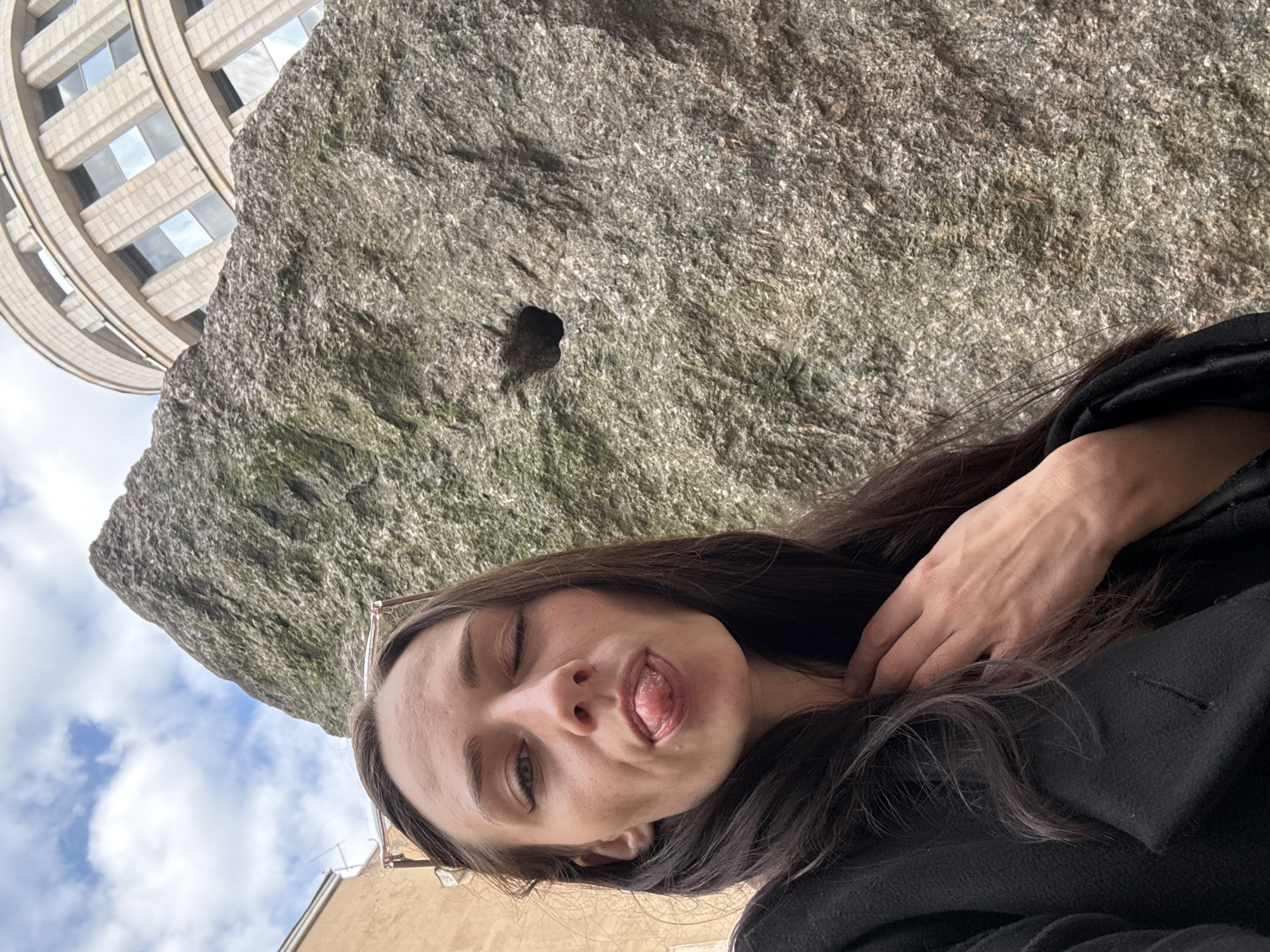 The image captures a playful outdoor selfie of a young woman in Saint Petersburg, Russia. In the foreground, a woman with long, dark hair and wearing a dark jacket winks at the camera while sticking out her tongue in a mischievous expression. Her right hand is visible near her neck and shoulder.
Behind her, a large, rough-textured grey rock dominates the mid-ground. The rock displays a mottled surface of various grey tones, hinting at its natural or aged state, and features a small, dark, irregular hole. In the background, a distinctive building with a curved, multi-tiered facade and reflective rectangular windows can be seen, partially obscured by the rock. The sky is visible above, characterized by white clouds against a light blue backdrop, indicating daytime. A glimpse of another building with a light brown wall is present in the lower right, grounding the scene in an urban environment. No text is visible in the image. The overall impression is casual and lighthearted.