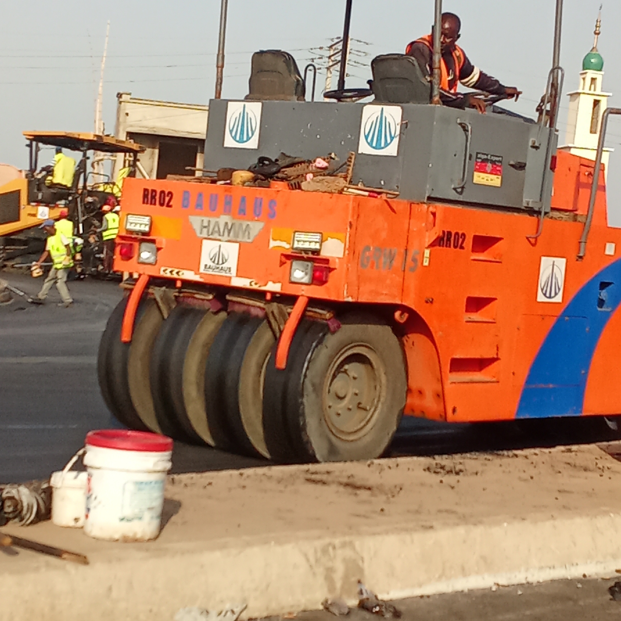 A wide shot shows a road paving construction project in Akwanga, Nigeria. An orange HAMM compactor with "RR02 BAUHAUS" in blue text on the back sits on a concrete road curb. The compactor is manned by a Black driver wearing an orange safety vest. The compactor has four rear tires and two seats. To the left of the compactor, a yellow and black paving machine, is surrounded by workers in yellow safety vests. A white building with a green-domed minaret is in the background. Two paint buckets are on the curb to the left of the compactor.
