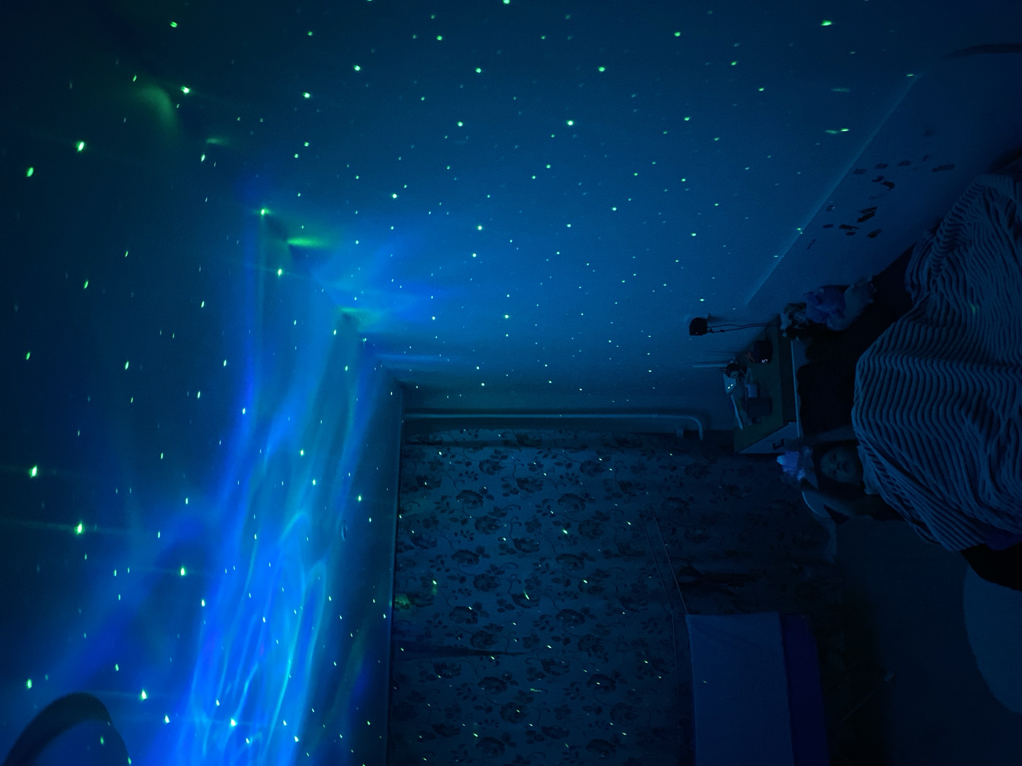 The image depicts a room illuminated by a star projector. The projection covers the ceiling and wall on the left with blue and green light. The wall displays a nebula effect with stars overlaid. A different wall is covered in floral wallpaper. A bed with striped bedding is present. A person is visible in the bed, lying down. A shelf with various items is partially visible next to the bed. The location is Chaikovsky, Russia.