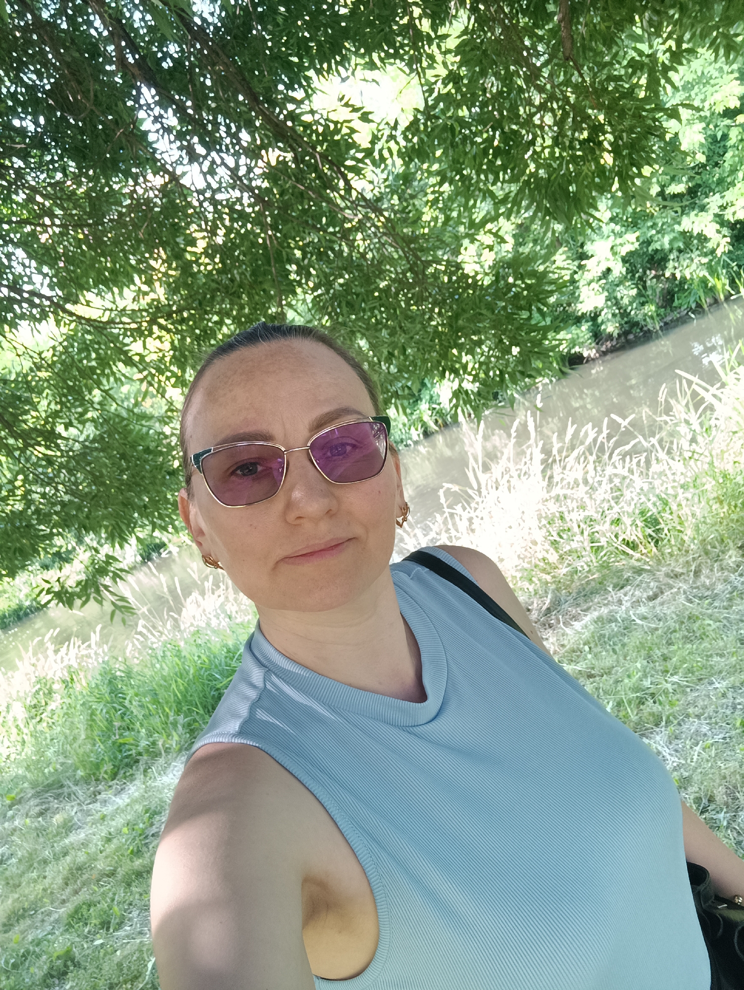 A woman wearing a light blue sleeveless top and eyeglasses is taking a selfie outdoors. She is positioned in the foreground, with the lower branches of a tree overhead and dappled sunlight filtering through the leaves. Behind her, a body of water, likely a river or canal, is visible, bordered by tall grass and vegetation. The setting appears to be a park or natural area, with the woman situated on a grassy bank beside the water. The overall impression is a casual, outdoor portrait.