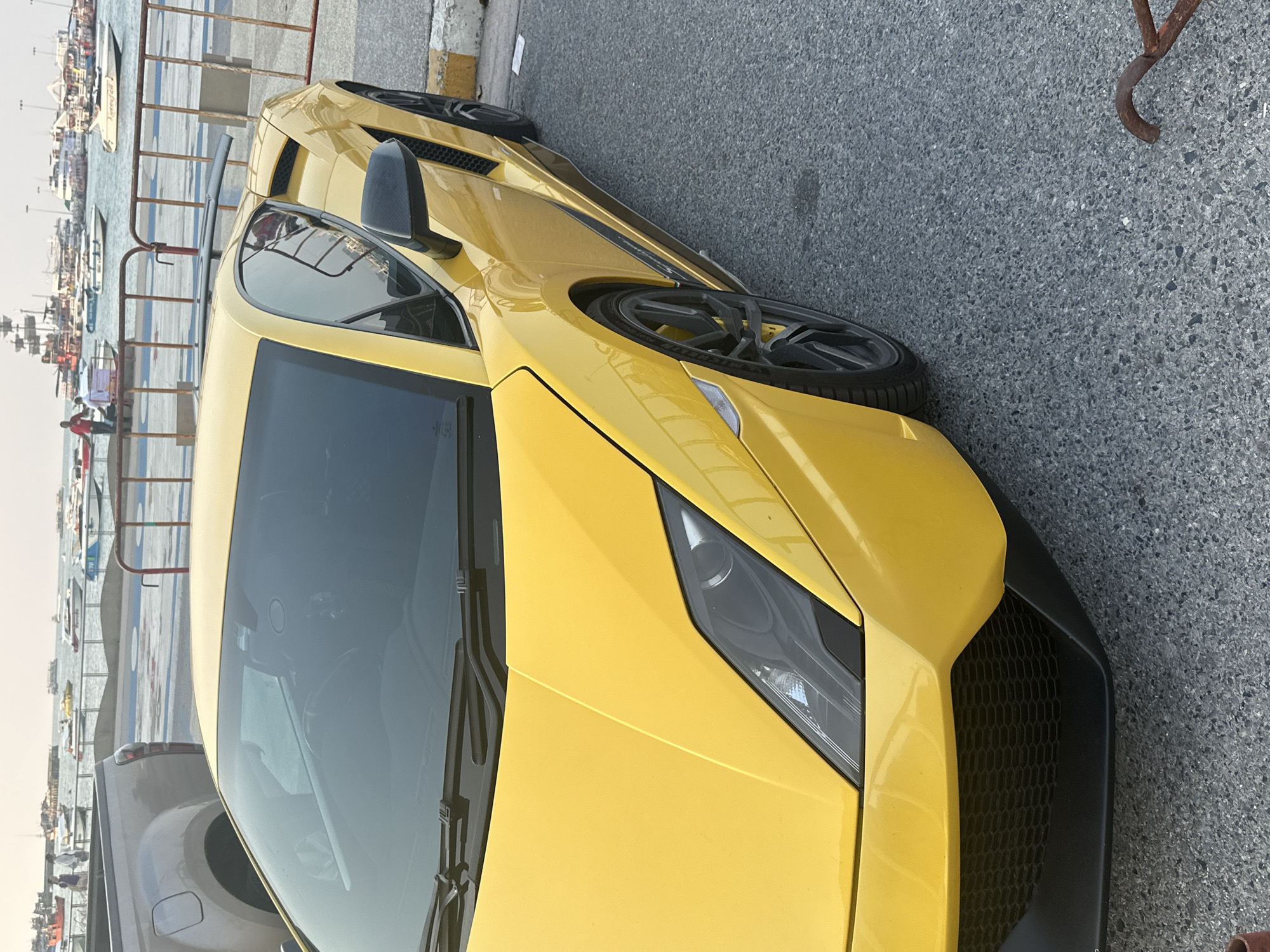 The image shows a yellow Lamborghini parked in Pattaya City, Thailand. The car is positioned near a waterfront area with railings and boats visible in the background. The car is angled towards the viewer, highlighting its front end and driver's side. The environment consists of the waterfront scene and paved ground where the car is parked.