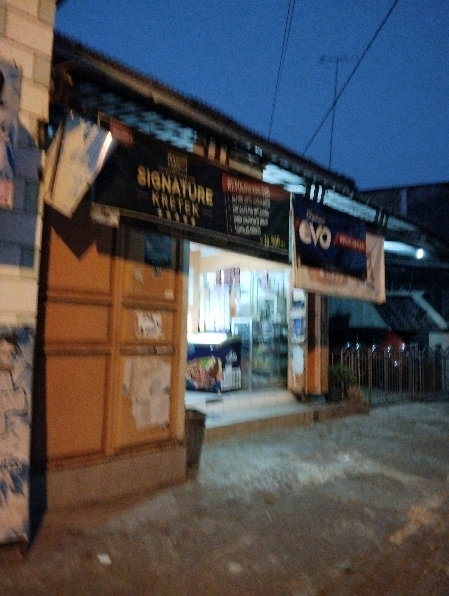 The image depicts the exterior of a store in Salatiga, Indonesia, during twilight hours. The store has a wooden facade with orange-colored doors and panels. Above the entrance, a dark sign reads "SIGNATURE KRETEK" in yellow, indicating a brand of cigarettes. To the right, a blue banner displays "EVO" in large white letters. The store's interior is visible through the open entrance, revealing shelves stocked with various products. A metal fence is visible in the background, along with glimpses of parked vehicles. The ground in front of the store appears to be paved with concrete or asphalt. The overall ambiance is that of a local retail establishment in an urban or semi-urban setting.