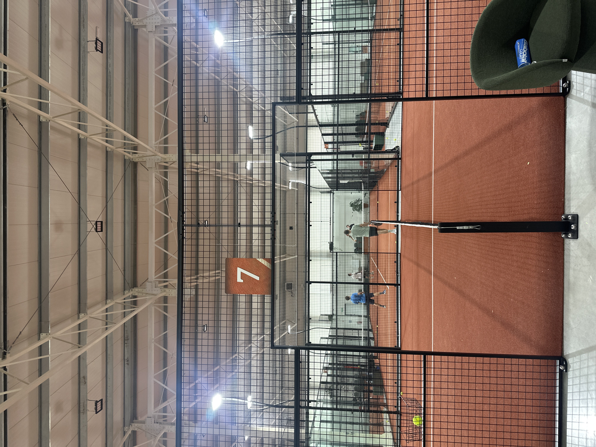 The image depicts an indoor padel tennis court, bathed in artificial light from overhead fixtures. The court itself has a reddish-brown playing surface marked with white lines, and is enclosed by black metal mesh fencing and glass panels. A black net spans the center of the court.
Three people are visible playing padel. On the far side of the net, one player wears a light-colored shirt and dark shorts, holding a racket. Another player, dressed in a blue shirt and dark shorts, is on the near side of the net, also with a racket. A third person in a light shirt is partly visible on the far side. Yellow padel balls are scattered on the court and contained within a wire basket in the foreground.
The setting features a high ceiling with exposed white metal trusses and beams, supporting light-colored wall panels. A prominent orange sign with the number "7" hangs from the ceiling, likely indicating the court number. In the immediate foreground, outside the court, a dark green armchair holds a blue can with the text "Babolat COURT" visible. Through the transparent barriers, glimpses of other padel courts and their players can be seen in the background, suggesting a multi-court indoor sports facility. The scene is well-lit, implying a play session occurring under artificial illumination, with no indication of outdoor weather or specific time of day.