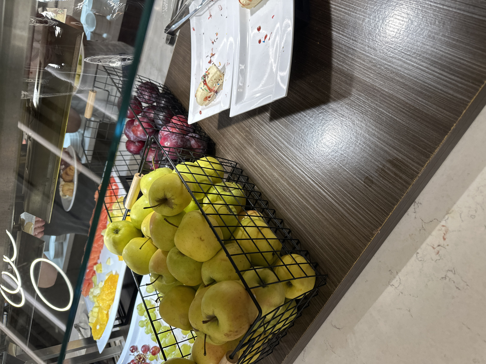 The image shows a buffet display with various food items in an indoor setting. There are several baskets filled with fruit including green apples and plums. Next to the fruit are white plates with remnants of food on them. The buffet display has an open section on the left showcasing slices of oranges. The food is placed on a dark wood countertop. The location is not discernible from the image, and therefore the city of Kazanlak, Bulgaria cannot be confirmed.