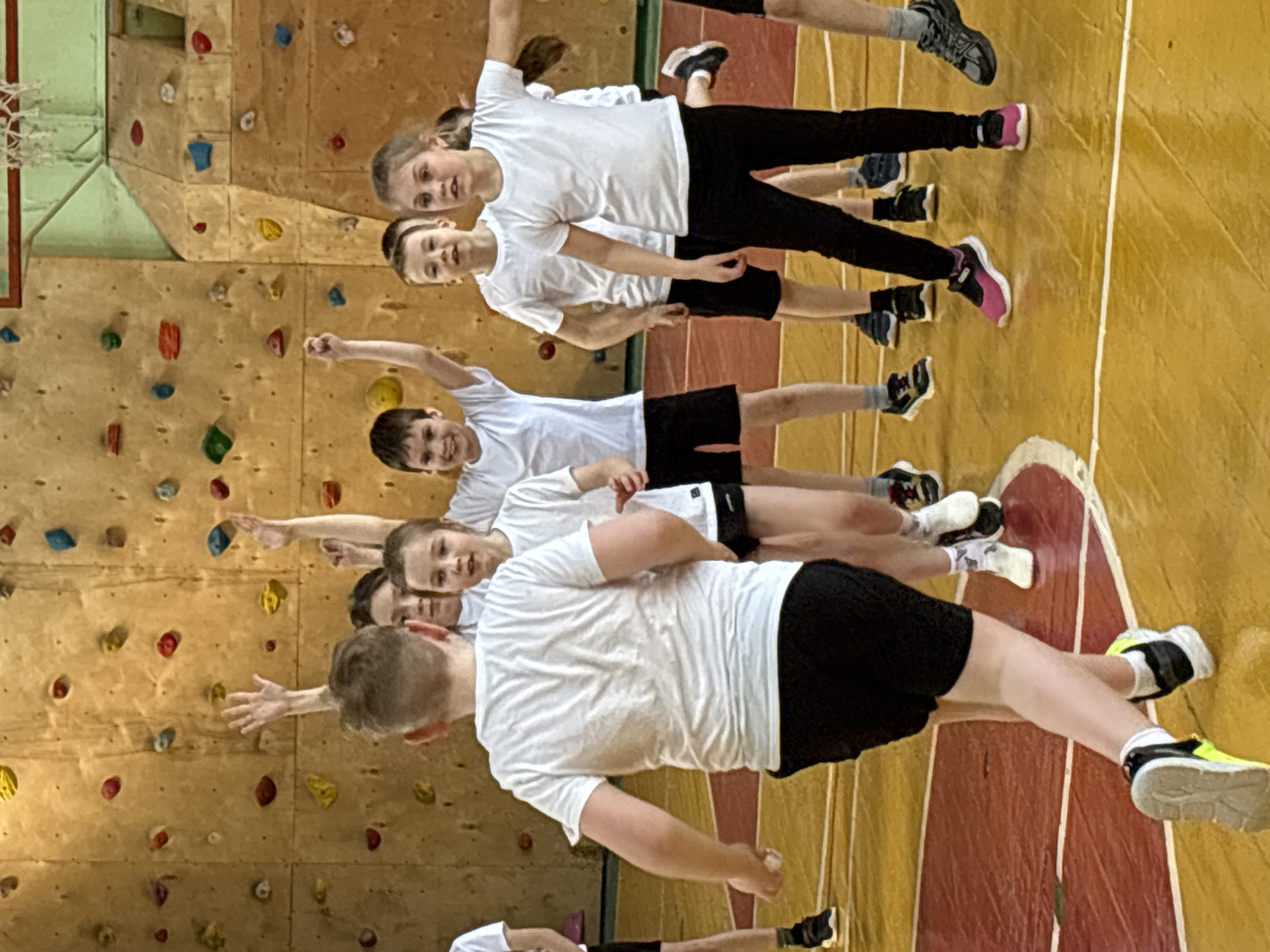 A group of approximately seven children are pictured indoors, likely in a gymnasium or sports hall in Izhevsk, Russia. The children, appearing to be in elementary or middle school, are lined up in a row, facing towards the viewer and slightly to the right, with their bodies oriented horizontally due to the image's rotation. All are wearing white t-shirts; some are in black shorts, while others wear black or dark-colored long pants.
The children seem to be participating in a physical activity or posing for a group photo. Many have their arms raised or extended, and most show smiling or engaged expressions, indicating enjoyment or anticipation.
The background features a large climbing wall constructed from wooden panels, adorned with numerous colorful climbing holds in shades of red, blue, yellow, green, and orange. In the upper left corner, part of a basketball hoop and backboard is visible. The floor is a light brown or yellow sports surface with red and white markings, consistent with a gym or sports court. The lighting is bright and even, suggesting an indoor setting during the daytime. No visible text is present in the image.