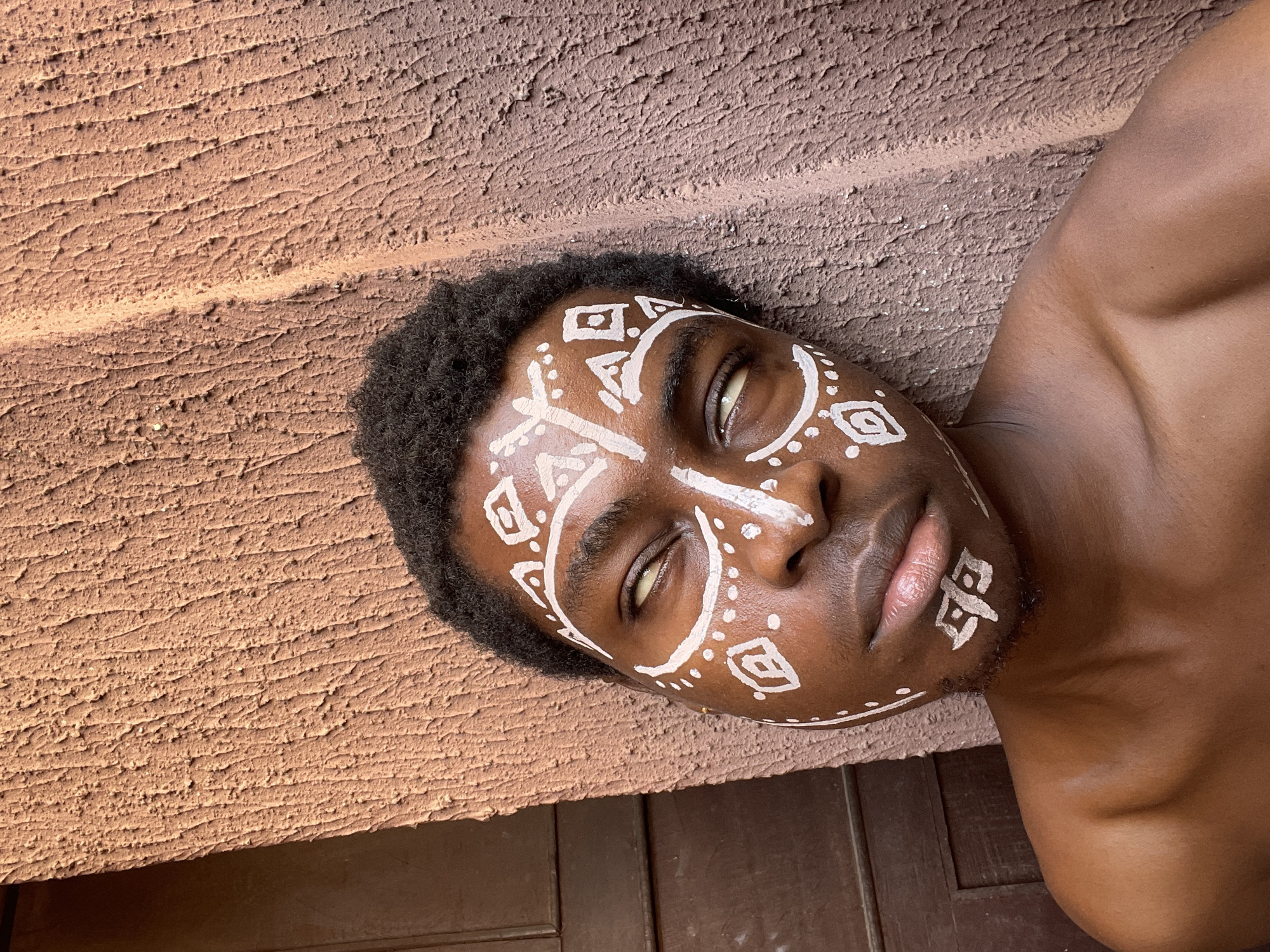 The image shows a dark-skinned person with tribal face paint lying near an exterior wall and a wood surface. The wall has a rough, textured, reddish-brown finish. The face paint is white, consisting of geometric shapes and dots. The person's head is tilted to the side, and the eyes are partially open. The wood surface appears to be a door or a frame. The location is identified as Nkpor, Nigeria.