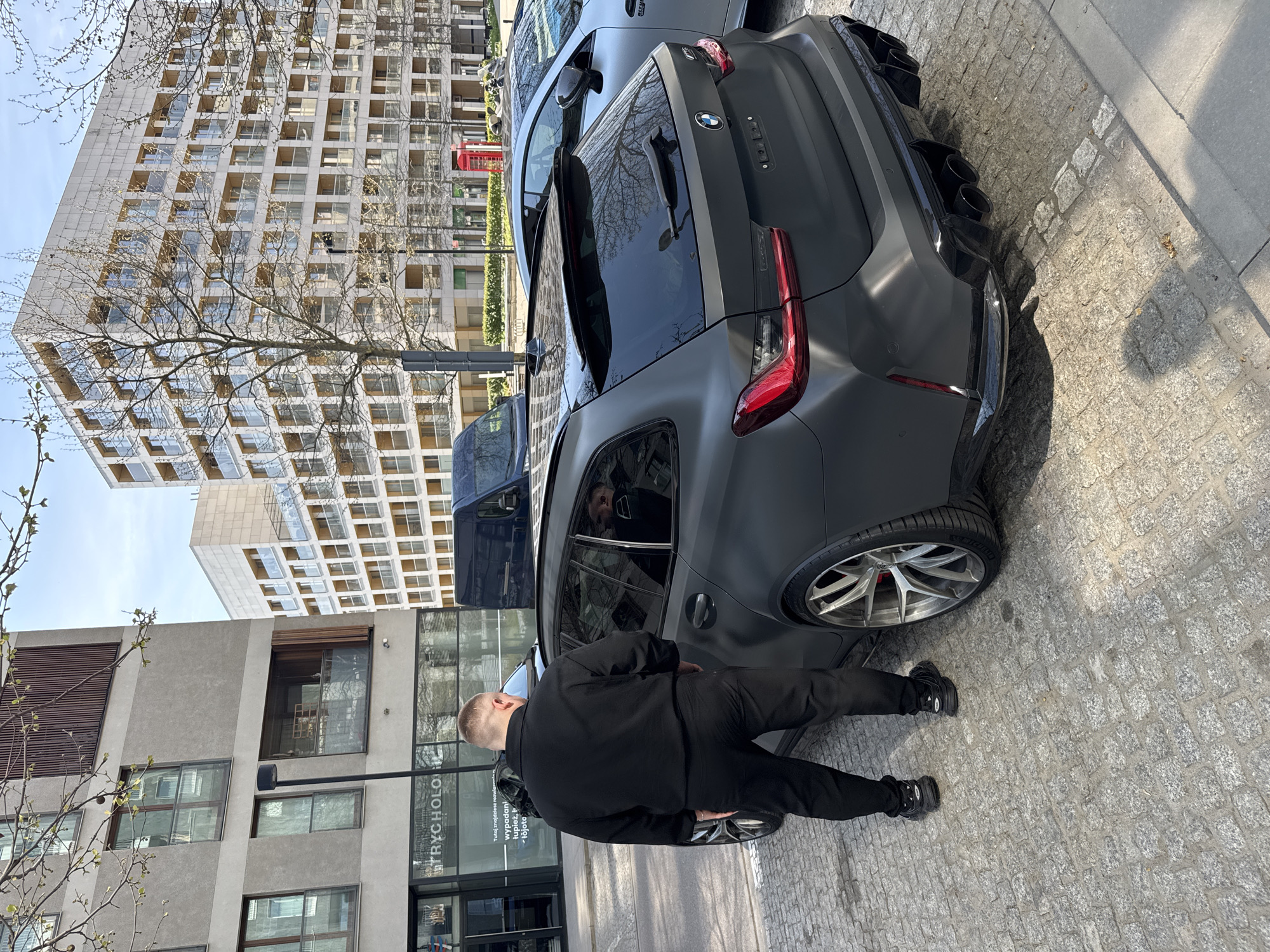A daytime shot shows a man in all black clothing getting into a matte grey BMW M3 Touring parked on a cobblestone street in Warsaw, Poland. The car is parked near a building with the sign "TRYCHOLOG" visible. In the background is another similar car, a red telephone box, and a tall apartment building with a grid-like facade. A bare tree branch is partially visible in the foreground.