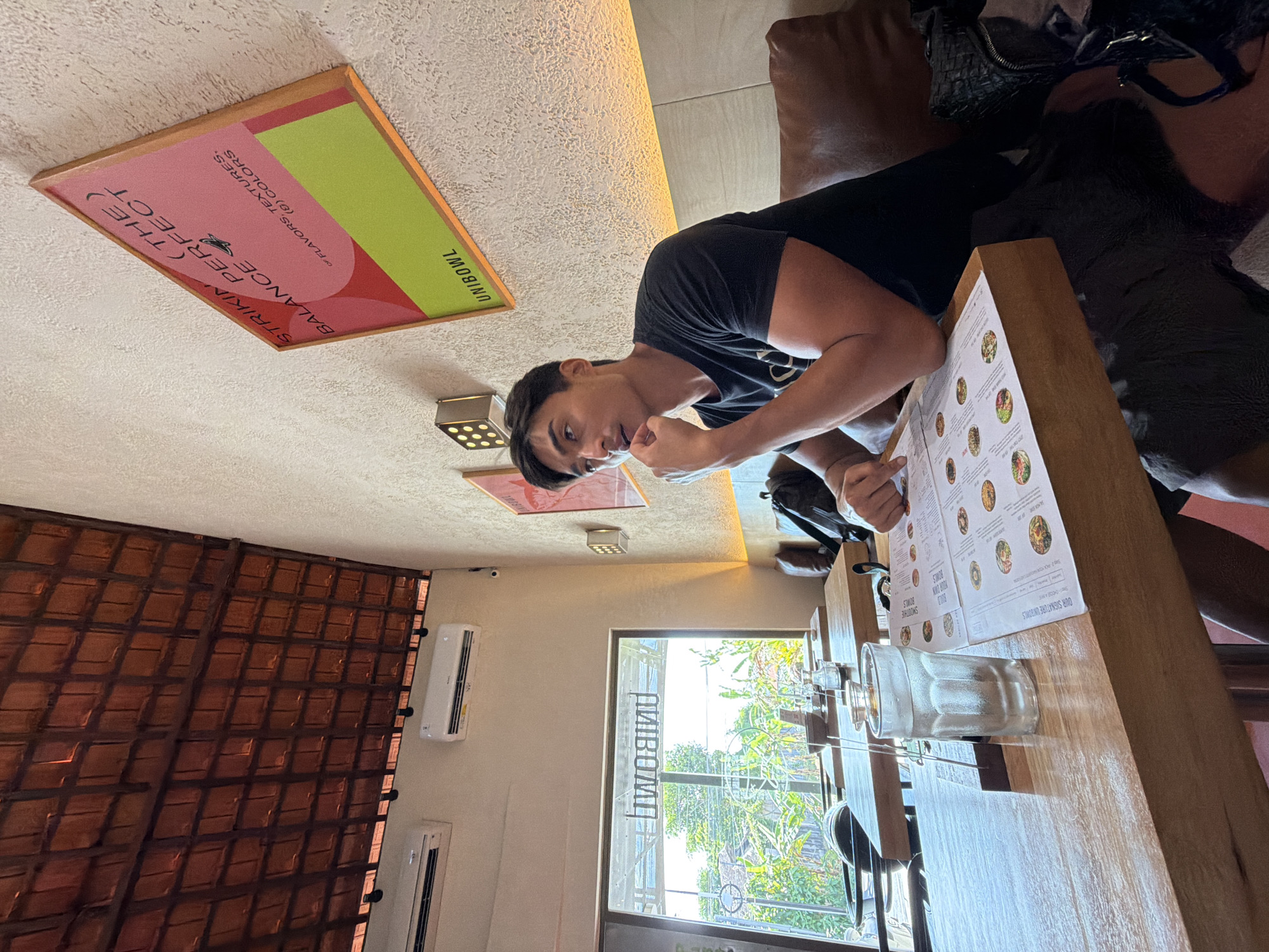 A medium shot shows a man reading a menu inside the restaurant “UNIBOWL,” located in North Kuta, Indonesia. The man is sitting at a wooden table with a glass of water. The menu in front of him has photos of the restaurant’s signature bowls. The restaurant’s name is written on the front door, and the restaurant's sign can be seen on the ceiling.