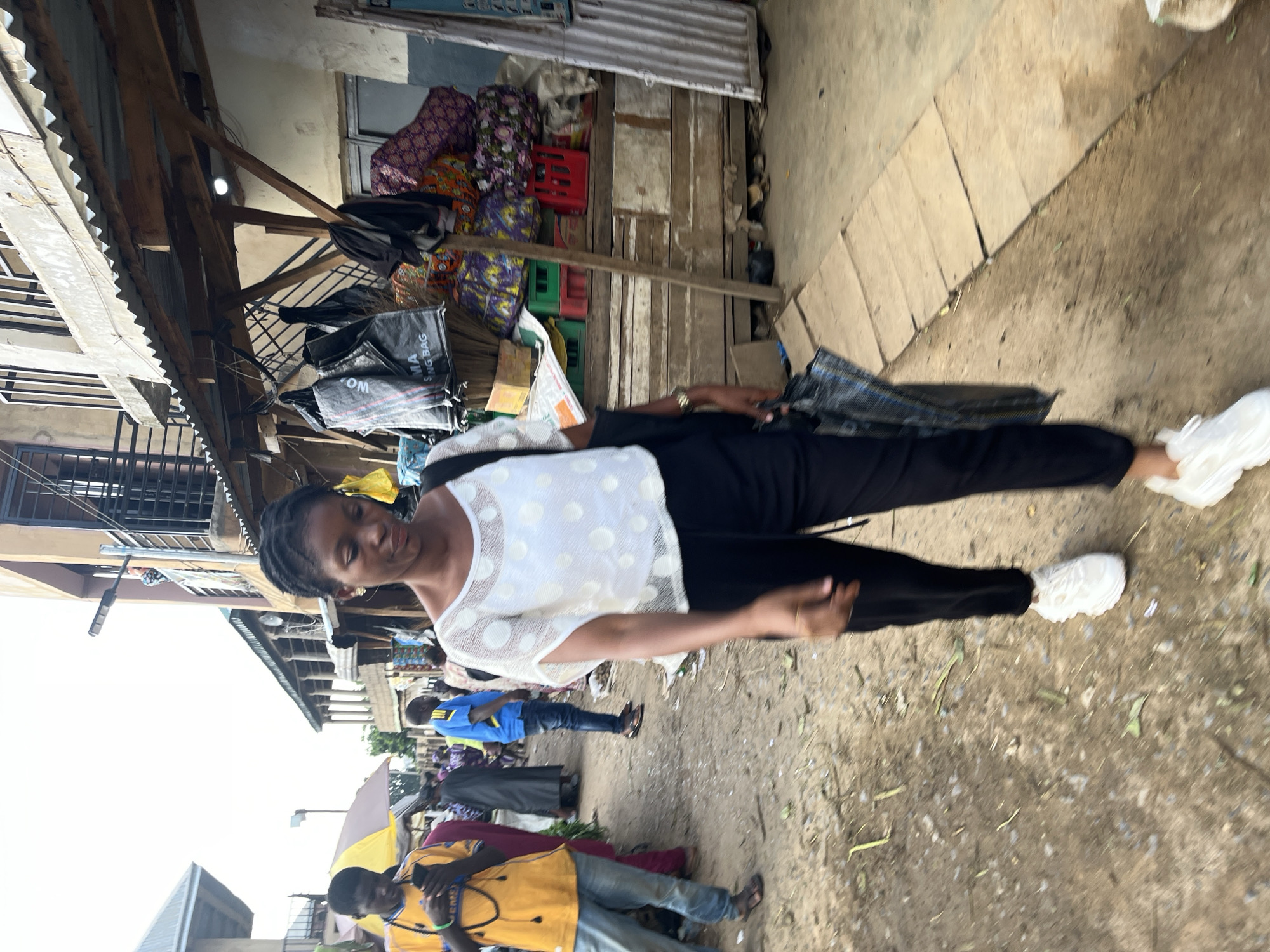 A young woman walks towards the camera on a dirt road in what appears to be an outdoor market area in Rafin Par, Nigeria. She is wearing a white, short-sleeved blouse with a circular pattern and black pants, along with white sneakers. She carries a black patterned bag in her right hand and a black tote bag over her left shoulder. Her gaze is directed forward and slightly downward. Behind her, other people are visible, some walking and others standing. To the right of the woman, stalls are set up with various items, including fabric, bags, and crates. The environment suggests a bustling local market with a mix of goods and people.