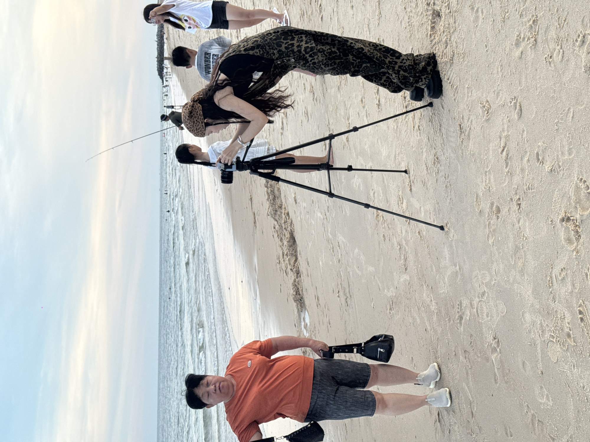 An outdoor, eye-level, full shot captures a sandy beach scene at Phu Thuy Ward, Vietnam, under a pale blue sky with scattered clouds. In the foreground, a woman with dark hair, wearing a leopard-print bandana and a matching jumpsuit, is focused on operating a DSLR camera mounted on a tripod. Beside her, a young girl in a white dress stands with her back to the camera. To the left, an adult with short dark hair and an orange t-shirt holds two black bags, one in each hand. Further back, other beachgoers are visible in the distance near the water, and a fishing rod stands upright on the sand. The ocean stretches across the background, with gentle waves reaching the shore.