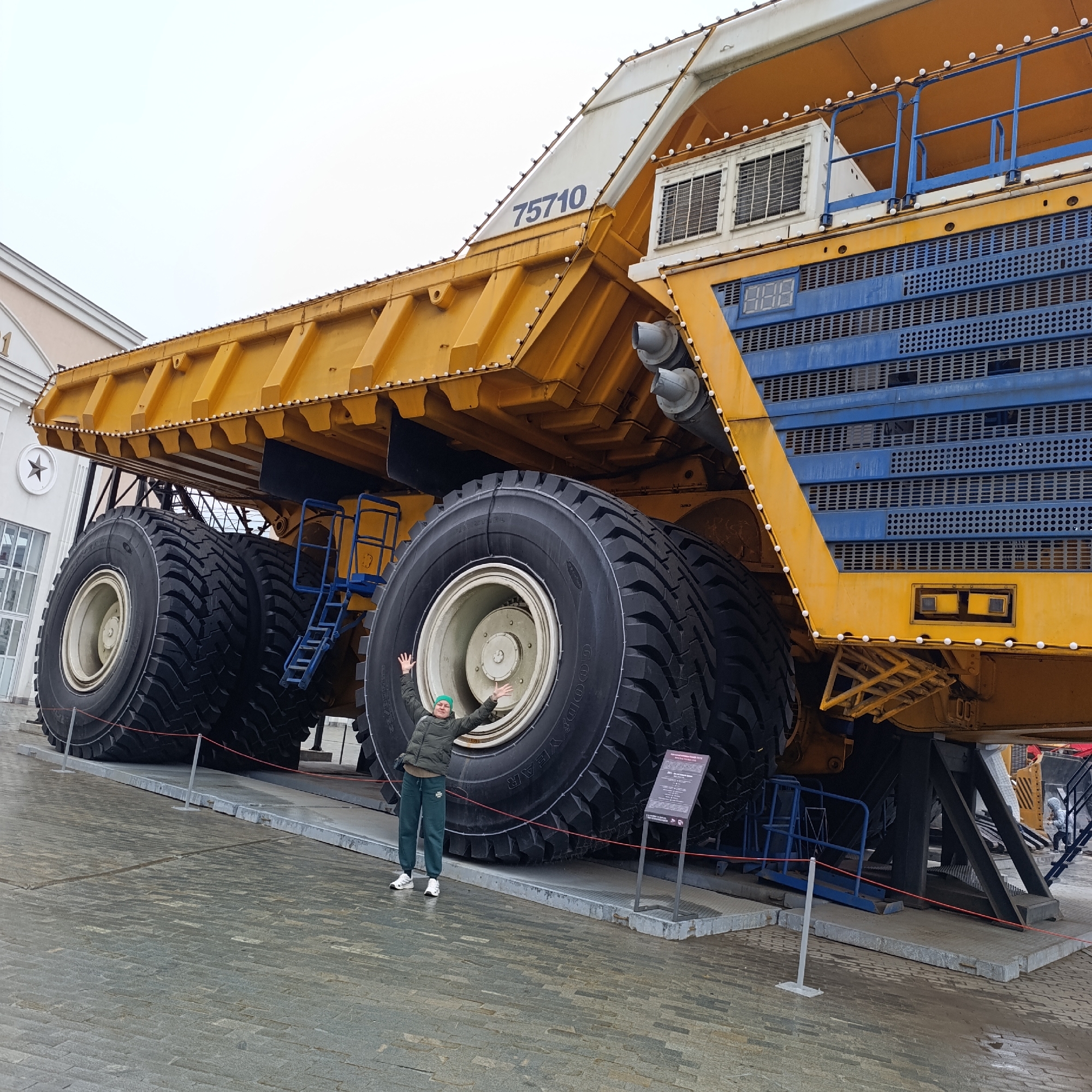 An outdoor, eye-level shot captures a person posing enthusiastically in front of a colossal yellow and blue mining dump truck, likely at an exhibition or museum in Verkhnyaya Pyshma, Russia. The vast scale of the truck is the dominant feature, with the person appearing tiny in comparison.
The massive dump truck is predominantly bright yellow on its body and dump bed, with distinctive blue grilles and panels on the front-facing side. Numerous small, white spherical lights adorn the edges of the truck's structure, possibly for display or nighttime illumination. The number "75710" is clearly visible on the side of the dump bed. Near the blue grilles, a digital display shows "888". Two large grey exhaust pipes extend from the upper part of the truck. Six enormous black tires are visible, with "GOODYEAR" and "100 YEAR" text faintly legible on the tire closest to the viewer. Blue metal ladders are affixed to the truck, indicating access points. The truck rests on a raised platform, with red ropes marking a barrier in the foreground.
The person, an adult, stands with arms outstretched and a joyful expression, dressed in a dark green puffer jacket, matching sweatpants, a light green beanie, and white sneakers. The ground is paved with grey paver stones and appears slightly wet. In the background, part of a light-colored building with "1" visible on its facade can be seen. The sky is overcast and bright, suggesting daytime with cloudy weather. A dark informational sign is positioned near the truck's front tire.