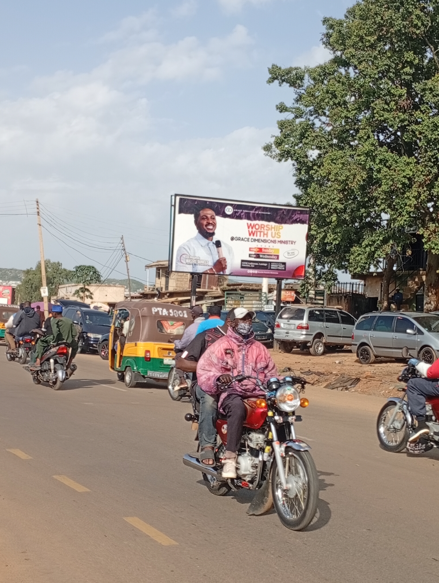 This is a photograph taken in the city of Rafin Par, Nigeria, showcasing a busy street scene. The image captures a vibrant procession of motorcycles and a tricycle (also known as a "Keke") navigating a paved road. In the foreground, two individuals on motorcycles are prominently featured, one wearing a light-colored jacket and a face mask, and another wearing a pink patterned jacket and black pants. Behind them, other motorcycles and the tricycle are visible, indicating a common mode of transportation in the area. A large billboard in the background advertises a church event, featuring a portrait of a man and the words "WORSHIP WITH US @ GRACE DIMENSIONS MINISTRY." The environment appears to be a typical urban setting in Nigeria, with buildings and trees lining the street. The sky is partly cloudy, suggesting a bright, possibly warm day. The overall impression is one of daily life and community activity in Rafin Par.