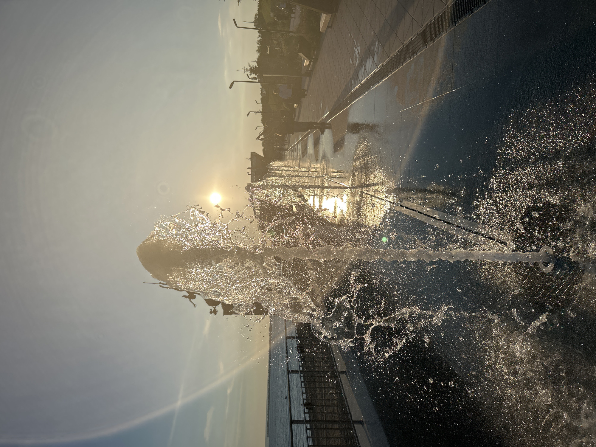 The image depicts a public fountain area with water jets spraying in the foreground. The sun is setting, casting a warm glow and creating reflections on the wet pavement. In the background, a walkway lines the fountain, with several people strolling and enjoying the evening. Beyond the walkway, there's a body of water, likely a bay or river, and a distant shoreline with trees. The scene appears to be the Embankment of Tolyatti, Russia, a popular recreational spot. The overall atmosphere is peaceful and idyllic, with the water and sunset creating a beautiful visual spectacle.