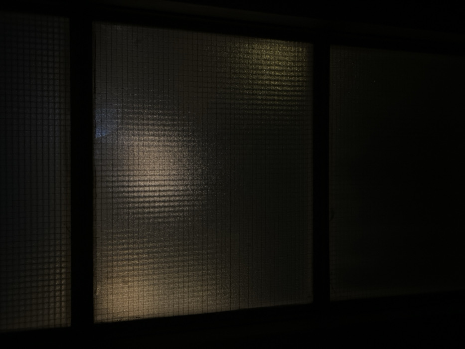 The image displays a close-up, low-light view of a multi-pane window located in Tel-Aviv, Israel. The window features patterned, translucent privacy glass, exhibiting a textured grid-like or ribbed surface that significantly obscures visibility of the exterior. Dark, rectilinear mullions separate the glass panes, forming the window frame. Through the patterned glass, several indistinct light sources are visible as blurred glows. Predominantly, a warm-toned light source is located in the central-left area of the main pane, with other fainter, yellowish light emanations visible through different sections, including towards the lower-right. No discernible human figures, specific identifiable objects, or distinct actions or interactions are visible within or beyond the obscured view provided by the translucent glass. The scene is characterized by deep shadow and ambient, diffused light originating from an external environment.