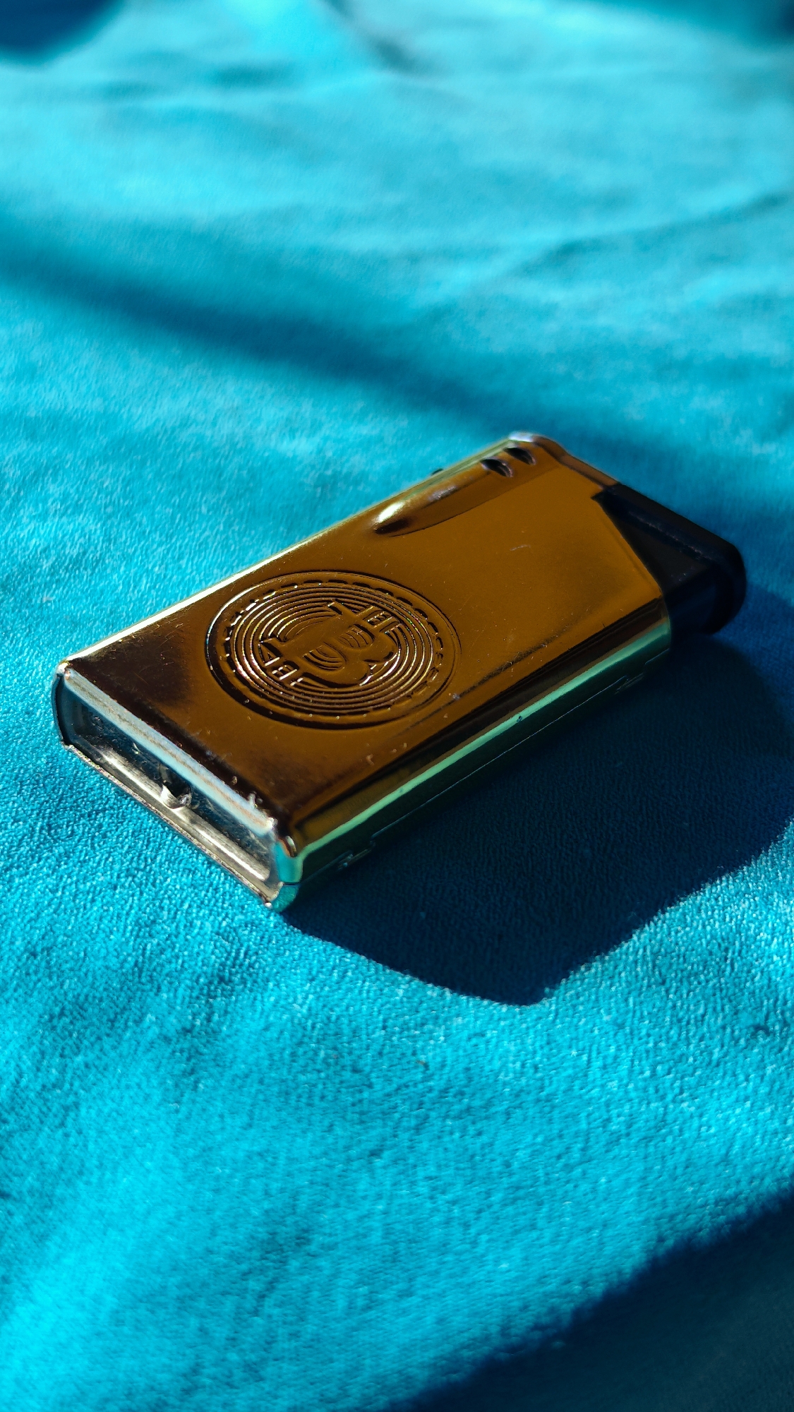 A gold-colored lighter with a Bitcoin logo embossed on its surface is positioned on a textured turquoise fabric. The fabric appears to be a towel or blanket, with visible fibers and a soft, plush texture. Sunlight casts a sharp shadow across the fabric and the lighter, indicating a strong light source from the side and slightly above. The lighter is rectangular with rounded edges and a black plastic component at one end. The Bitcoin logo is centrally located on the upper surface of the lighter, with intricate concentric circles and the Bitcoin symbol clearly visible. The overall scene is static, with no people or actions depicted. The image does not contain any specific geographical markers that would place it in Popovo, Bulgaria or any other city.